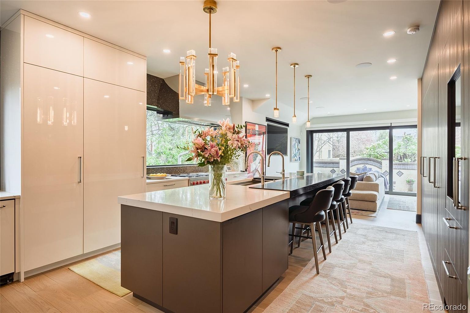 This is a modern kitchen with a large island featuring a white countertop and dark base, complemented by bar stools. The kitchen includes sleek, glossy cabinetry and modern lighting fixtures, including a chandelier and pendant lights. A large window provides natural light and a view to the outside, while the open layout connects to a living area, enhancing the sense of space.