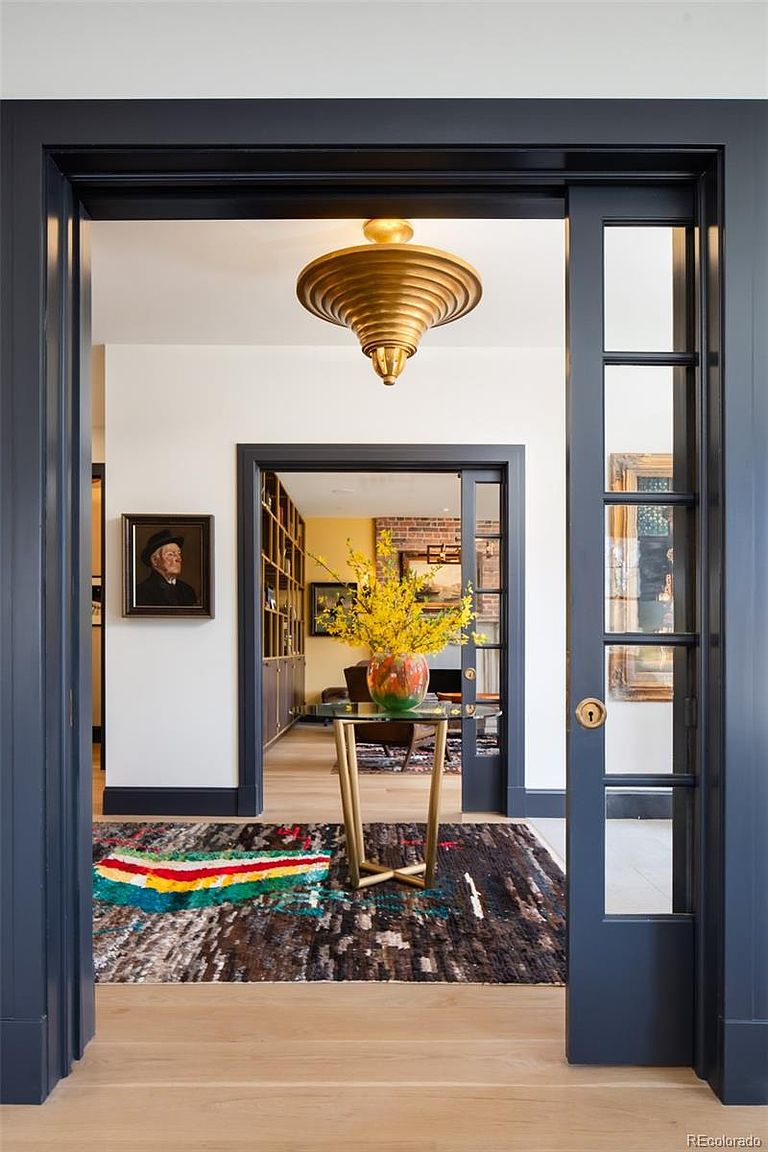 The image showcases an interior hallway with a view into another room, framed by dark blue door frames. A striking gold chandelier hangs from the ceiling, and a colorful rug adds a vibrant touch to the wooden floor. A glass-topped table with a vase of yellow flowers sits in the hallway, creating an inviting and stylish transition space.