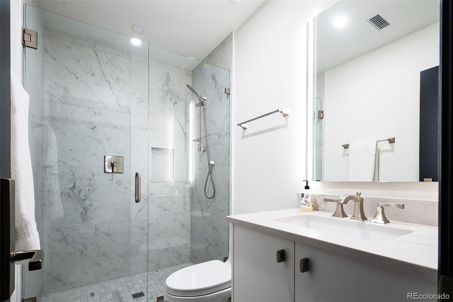 This is a modern bathroom featuring a glass-enclosed shower with marble tiling and a built-in niche. The vanity has a white countertop and cabinet with dark hardware, complemented by a sleek faucet and a well-lit mirror. The overall impression is clean, bright, and luxurious.