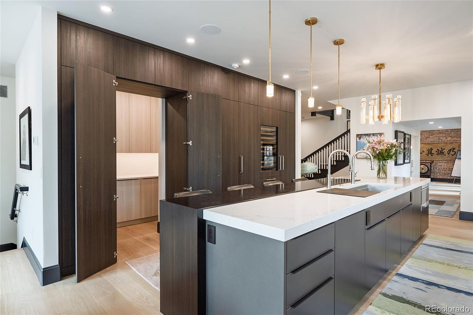 This is a modern kitchen with dark wood cabinetry and a large island featuring a white countertop and gray base. The kitchen includes pendant lighting, stainless steel appliances, and a hidden pantry. The overall impression is sleek and sophisticated, with a focus on clean lines and high-end finishes.