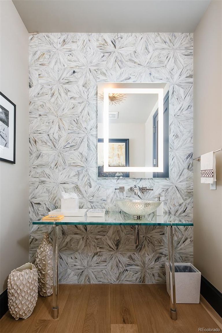 This is a stylish guest bathroom featuring a unique geometric tile wall that serves as a focal point. A modern, illuminated mirror hangs above a glass countertop vanity with a vessel sink. The space is well-lit and exudes a contemporary, upscale feel, making it a desirable feature for potential buyers.