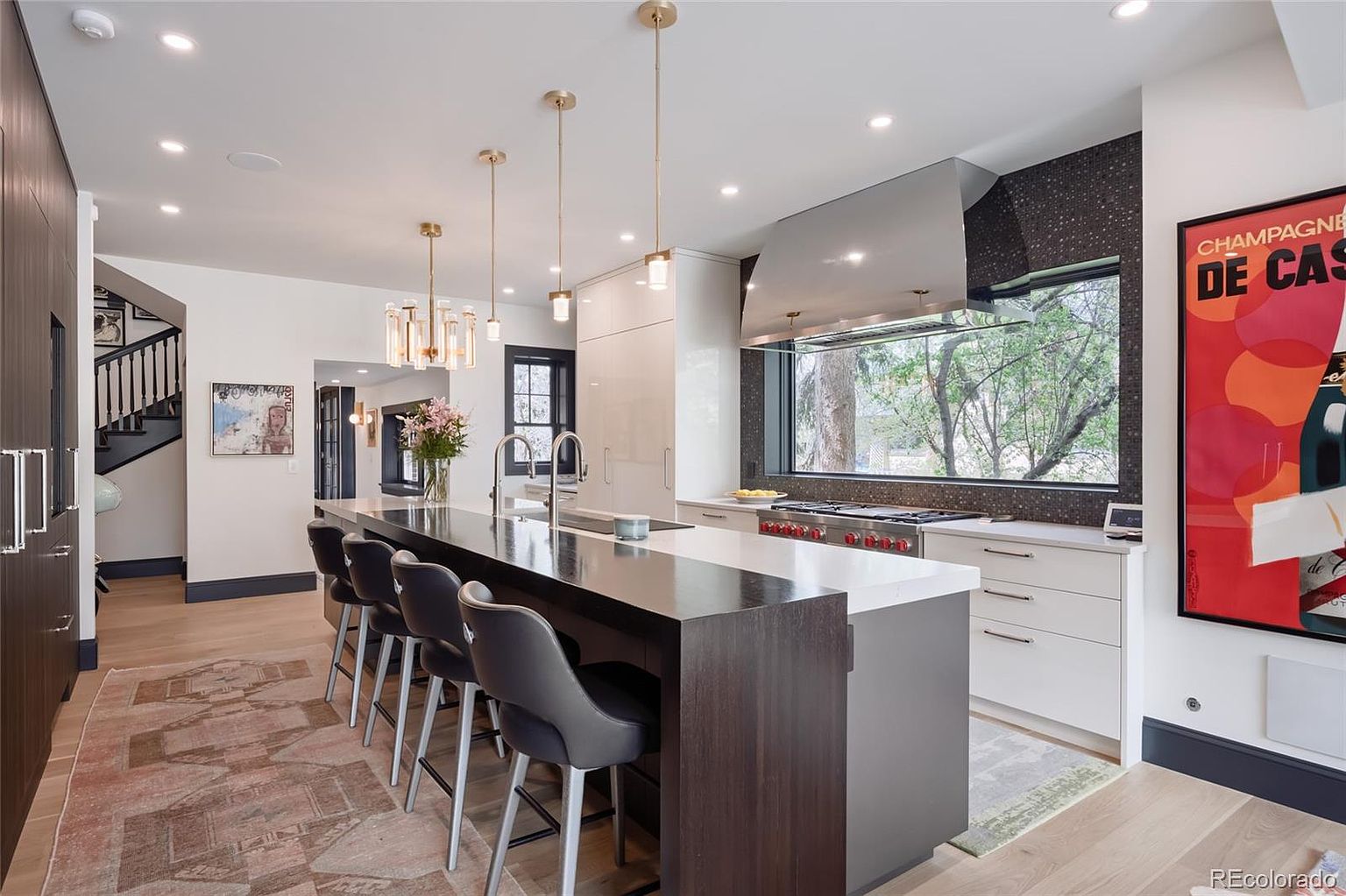 This is a modern kitchen with a large island featuring a dark wood base and a white countertop. The kitchen includes stainless steel appliances, white cabinetry, and a large window above the stove offering natural light. The space is illuminated by pendant lighting and recessed lighting, creating a bright and inviting atmosphere.