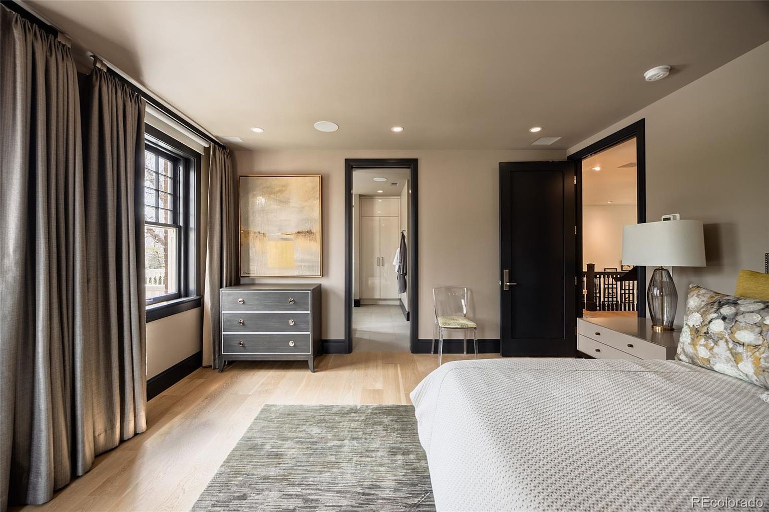 This is a primary bedroom featuring a neutral color palette with dark trim around the windows and doors. The room includes a dresser with artwork above it, a walk-in closet, and a view into another room, possibly a bathroom or sitting area. The flooring is light hardwood, and a patterned rug adds texture to the space.