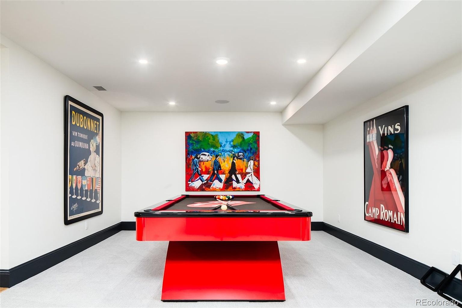 This is a well-lit basement featuring a red pool table as the focal point, positioned beneath a colorful art piece. The room is decorated with framed vintage posters on the walls, complemented by a neutral-toned carpet and dark trim. The space appears clean and modern, suitable for entertainment.