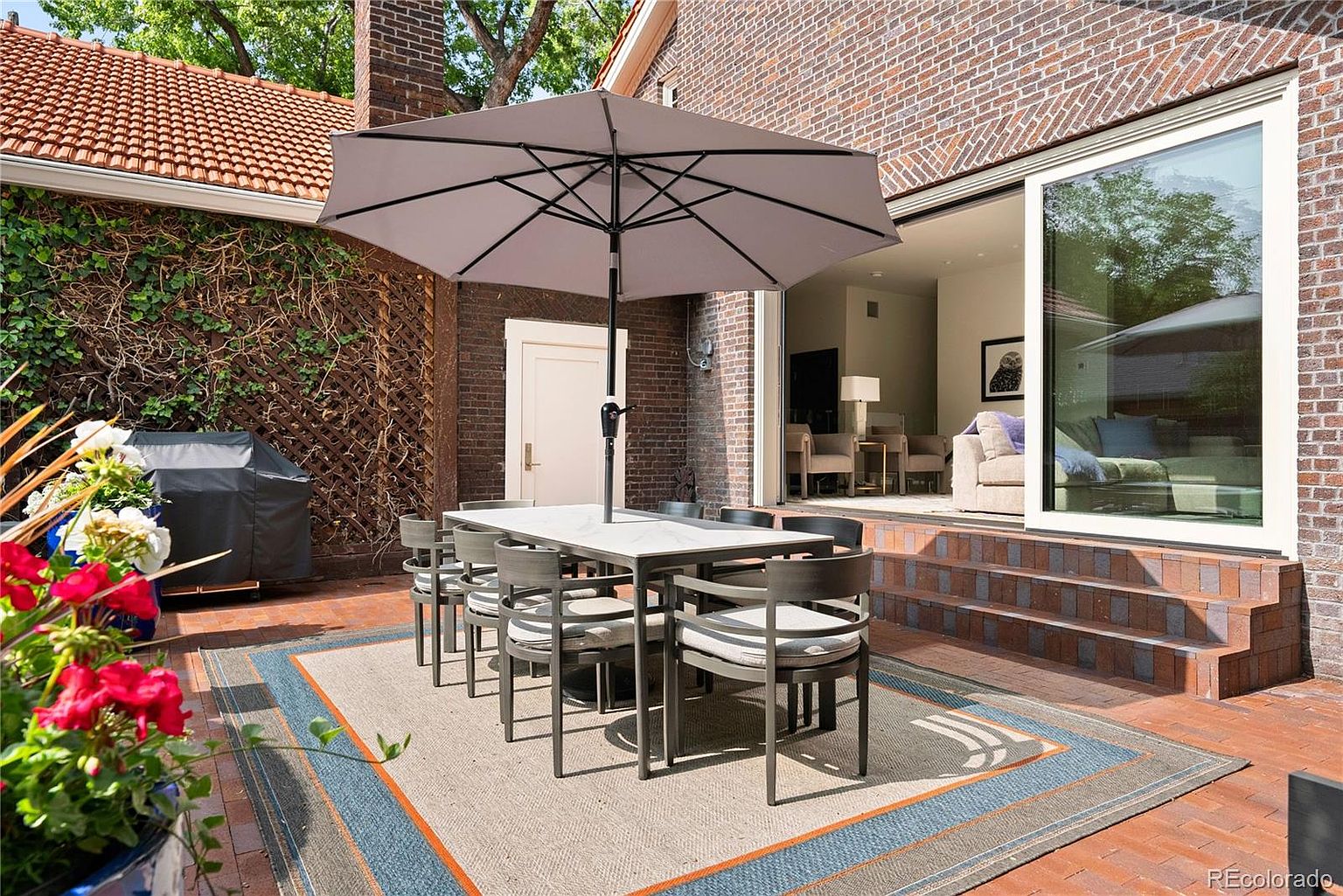 This is a well-staged outdoor patio area, perfect for entertaining. The space features a large dining table with seating for eight, shaded by a large umbrella. A stylish outdoor rug defines the space, and the patio is surrounded by brick walls, one covered in climbing vines, creating a private and inviting atmosphere.