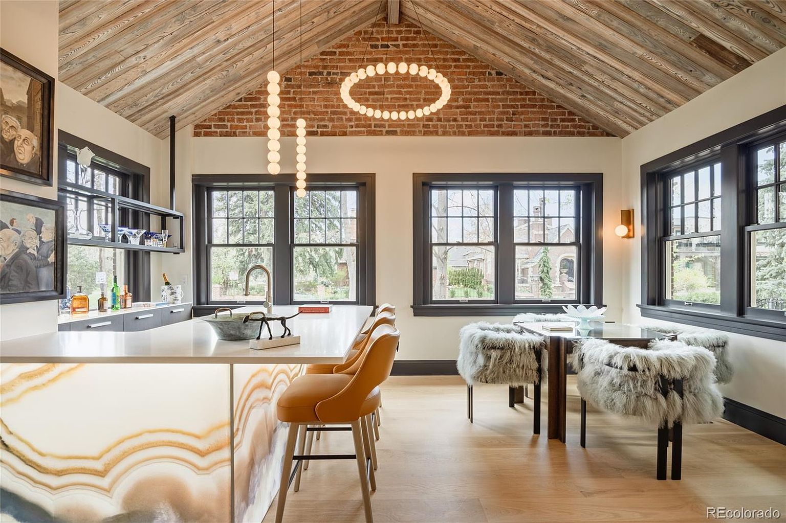 This interior shot showcases a modern kitchen with a unique design. The kitchen features a large island with a striking, patterned base, complemented by orange bar stools. The room is well-lit with natural light from the windows and modern pendant lighting, highlighting the wooden ceiling and brick accent wall.