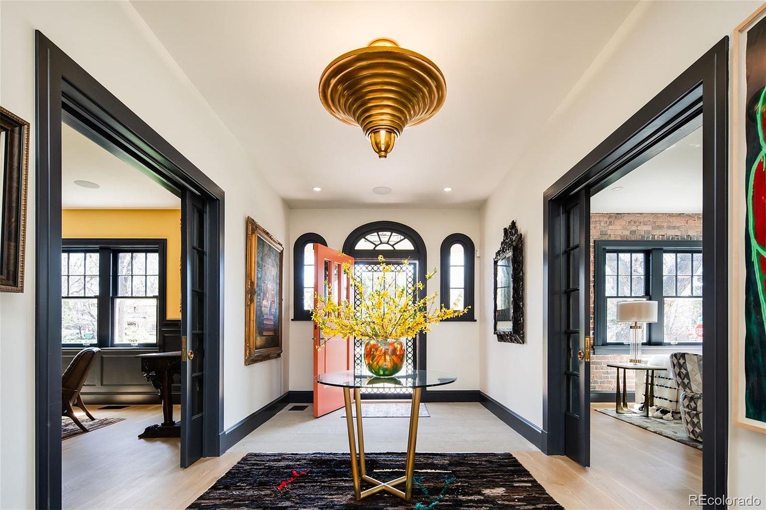 This is an interior shot of a grand hallway featuring a striking gold chandelier as a focal point. The hallway is flanked by doorways with dark trim, leading to other rooms, and a round glass table with a floral arrangement sits atop a patterned rug in the center. The walls are painted in a neutral tone, and the flooring is light wood, creating a bright and inviting space.