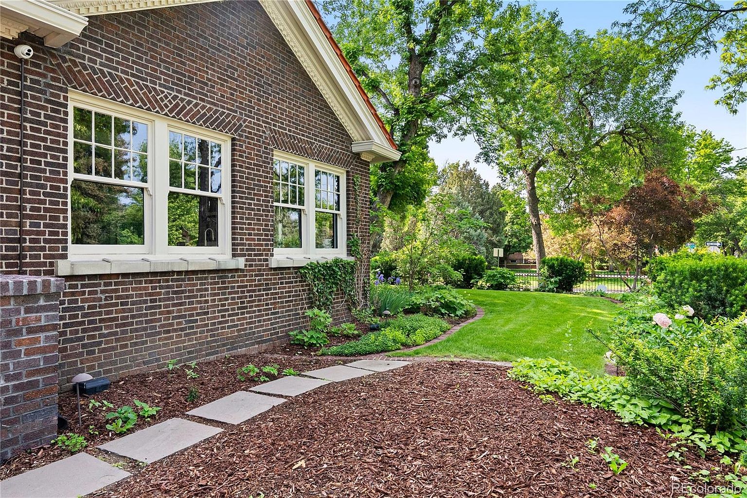 This image showcases the exterior of a brick house with a well-maintained yard and garden. A stone pathway leads through the mulched garden area, featuring various plants and greenery. The lawn is lush and green, with mature trees providing shade and enhancing the property's curb appeal.