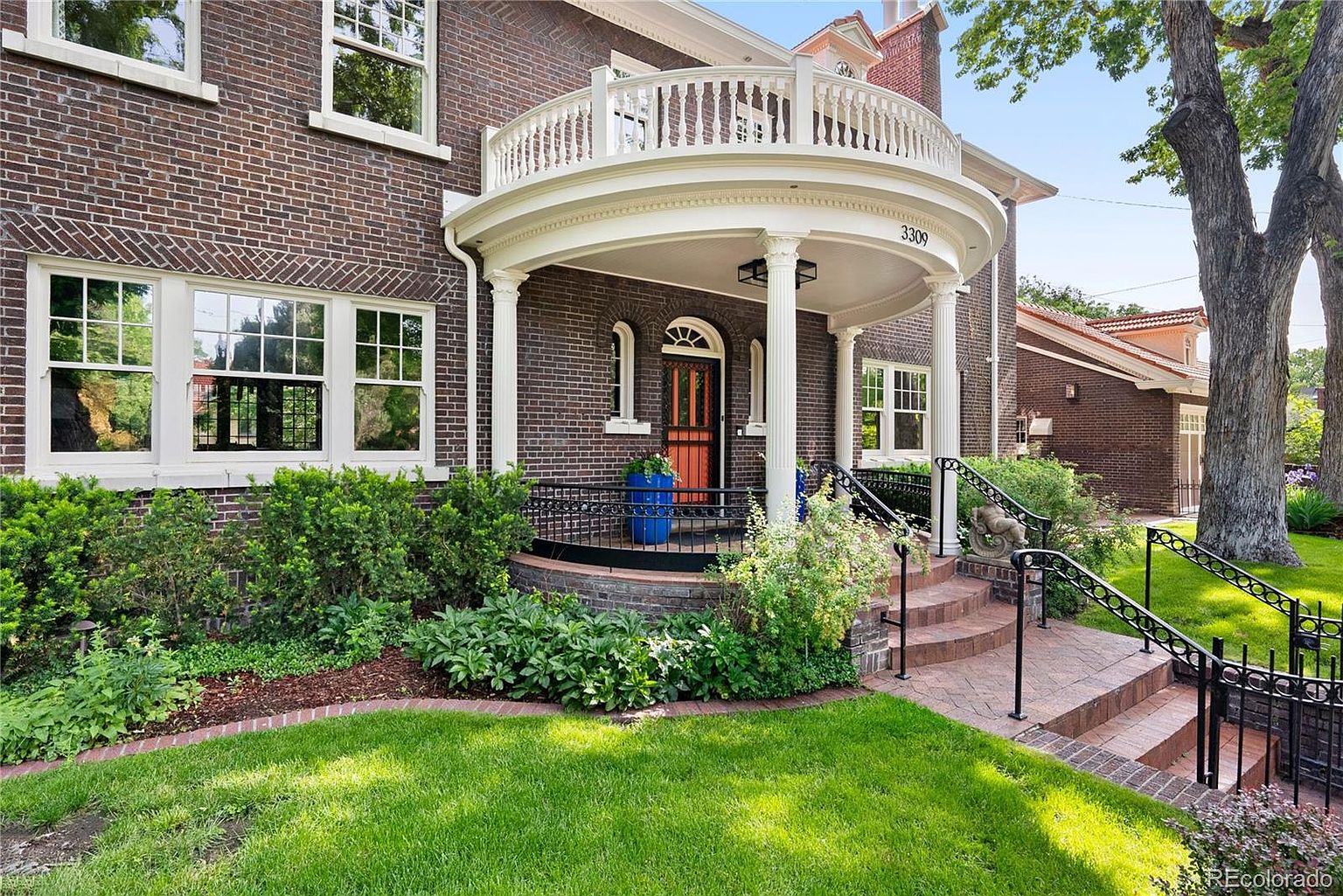 This is a front exterior view of a two-story brick home with a prominent curved balcony above the front porch. The house features white columns supporting the porch roof, a well-manicured lawn, and mature landscaping. The overall impression is one of classic elegance and curb appeal.