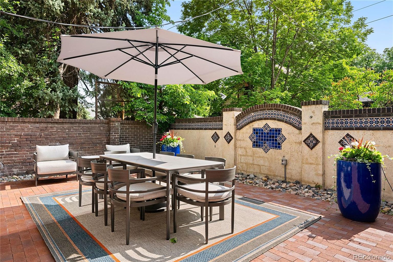 This image showcases a beautifully arranged outdoor patio area, perfect for entertaining or relaxing. The space features a dining table with chairs under a large umbrella, a comfortable armchair, and decorative planters. The patio is paved with brick and includes a stylish outdoor rug, creating a welcoming and sophisticated atmosphere.
