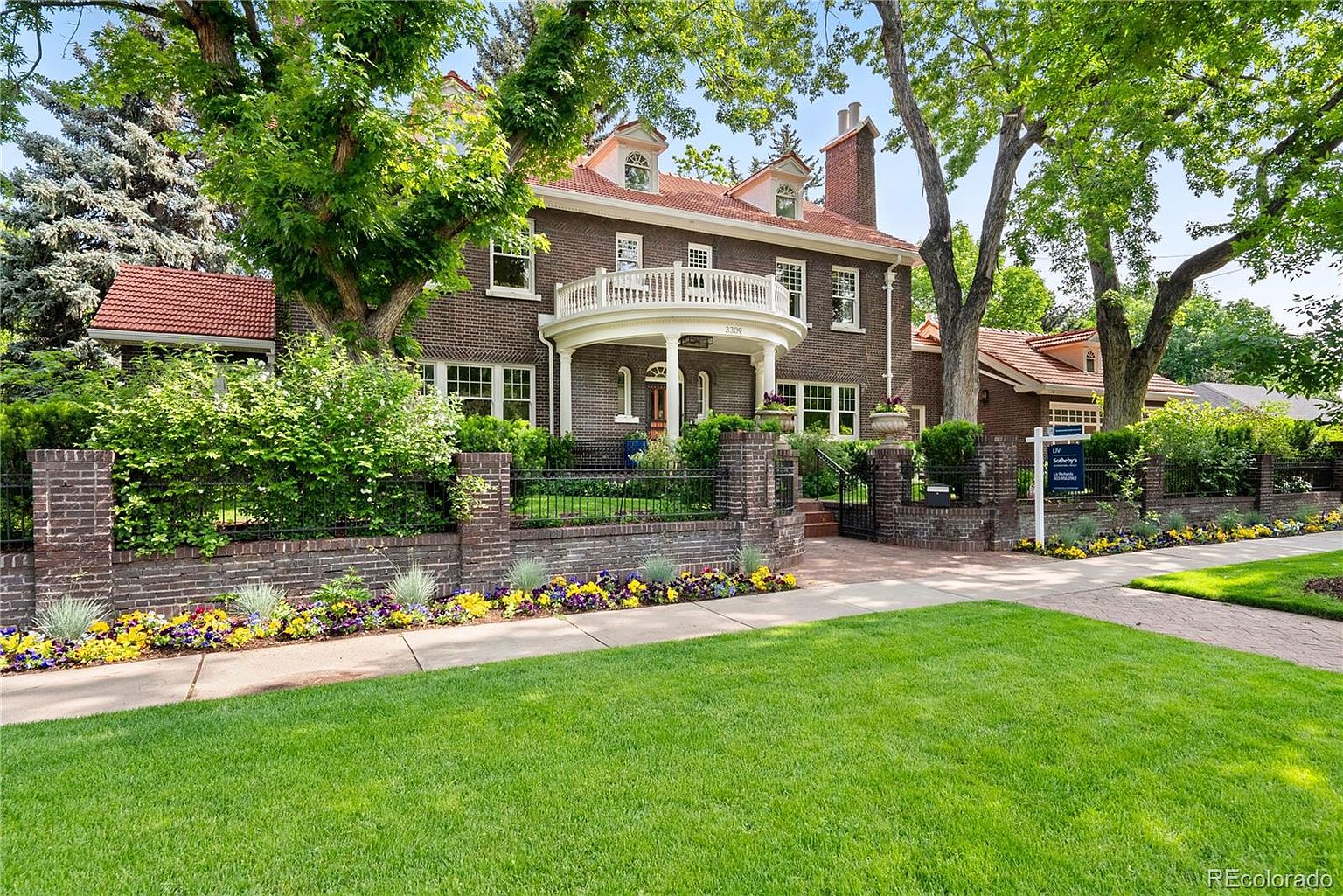 This is a front exterior view of a stately brick home with a red tile roof and a prominent white balcony. The property features a well-manicured lawn, mature trees, and a brick fence adorned with colorful flower beds. The overall impression is one of classic elegance and curb appeal.