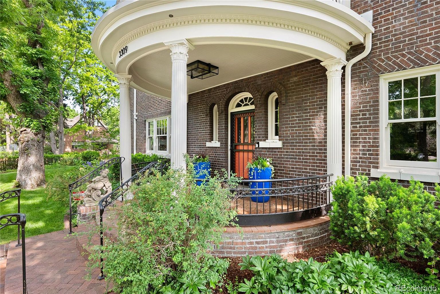 This is a charming entryway featuring a curved porch with white columns and a brick facade. The front door is a warm orange color, flanked by arched windows. Lush greenery surrounds the entrance, adding to the home's curb appeal and creating a welcoming atmosphere.