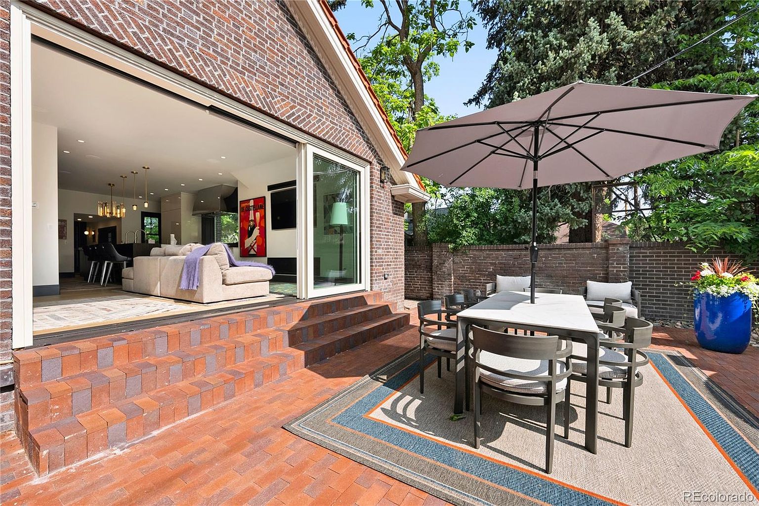 This image showcases a beautifully designed outdoor patio area, seamlessly blending indoor and outdoor living. The patio features brick flooring, a dining set with an umbrella, and lush greenery surrounding the space. Large sliding glass doors open into the home, creating an inviting and airy atmosphere.