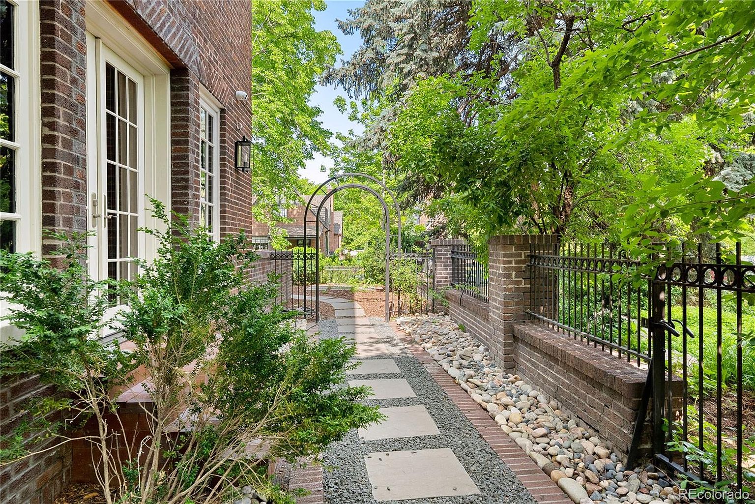 This image showcases a charming garden pathway leading through an arched gate. The pathway is constructed with stone pavers and gravel, bordered by brick walls and wrought iron fencing. Lush greenery and trees surround the area, creating a serene and inviting outdoor space.