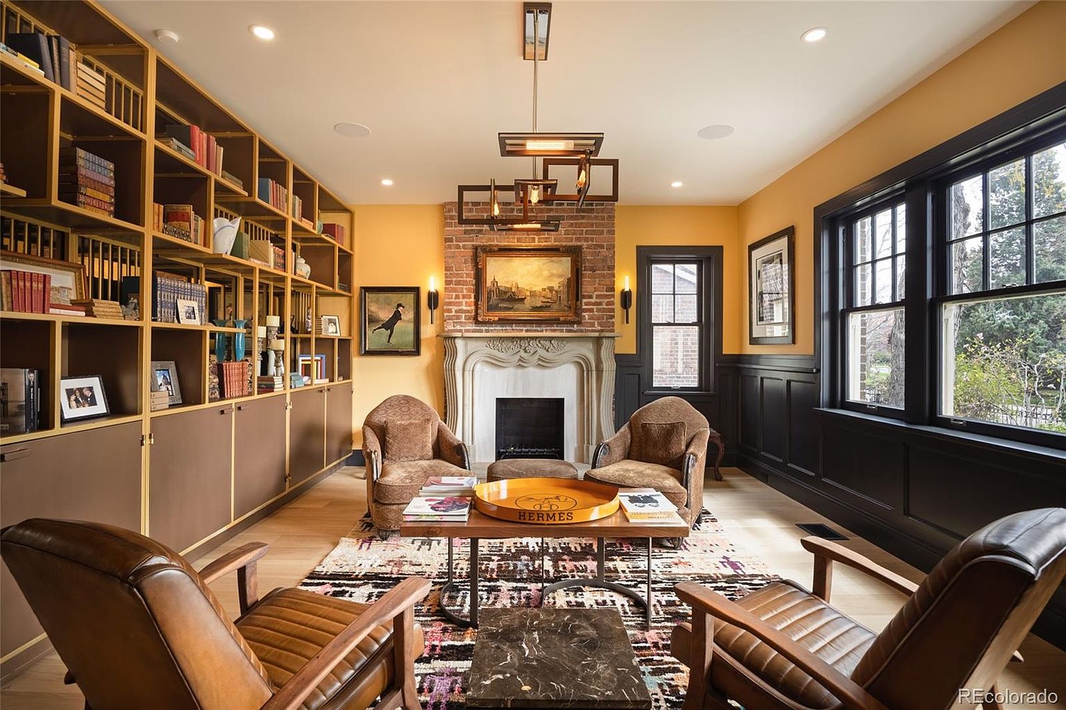 This is a cozy and sophisticated living room featuring a large built-in bookshelf, a fireplace with a decorative mantel, and comfortable seating arrangements. The room is decorated in warm tones with dark wood accents, creating a refined and inviting atmosphere. A patterned rug anchors the space, and natural light streams in through the windows.
