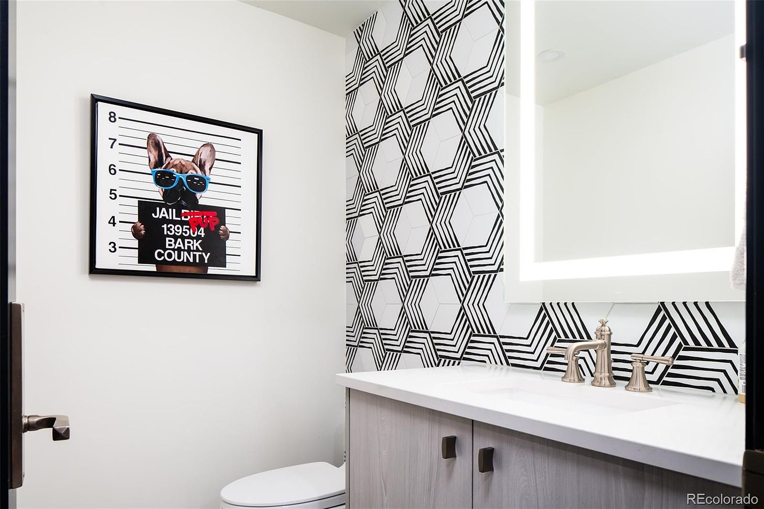 This is a modern guest bathroom featuring a geometric black and white tile accent wall, a light gray vanity with a white countertop, and a framed dog print. The space is well-lit and has a contemporary design, creating a stylish and inviting atmosphere. The perspective is from the doorway, showing the toilet, vanity, and accent wall.