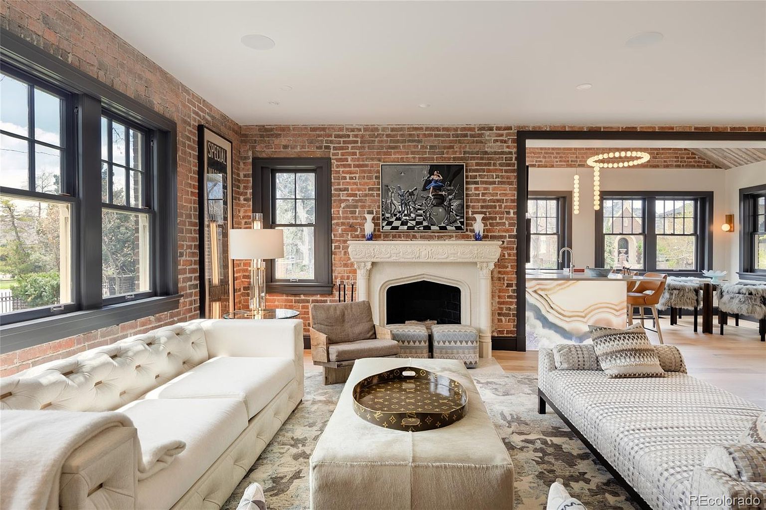 This is a beautifully designed living room featuring exposed brick walls and large windows that provide ample natural light. The room is furnished with a plush white sofa, a chaise lounge, and a large ottoman, creating a comfortable and inviting space. A fireplace serves as a focal point, and the open layout seamlessly connects to the kitchen area, enhancing the home's flow and functionality.