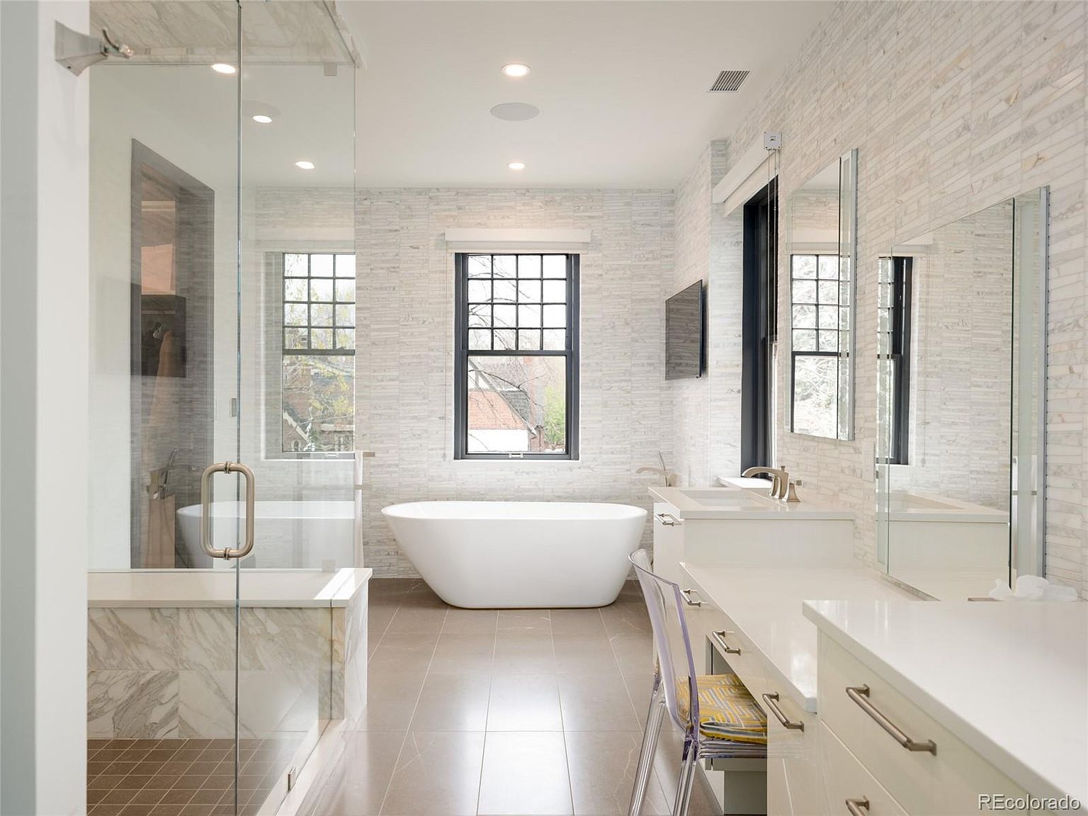 This is a luxurious primary bathroom featuring a freestanding soaking tub positioned beneath a window, a glass-enclosed shower, and a double vanity with ample counter space. The walls are adorned with light-colored, textured tiles, and the flooring is a neutral tone. The overall impression is one of spa-like tranquility and modern elegance.