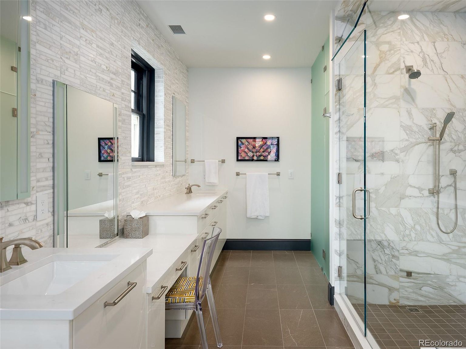 This is a primary bathroom featuring a double vanity with white countertops and cabinetry, complemented by brushed nickel fixtures. A large, glass-enclosed shower with marble tiling adds a luxurious touch. The room is well-lit with recessed lighting and natural light from a window, creating a bright and inviting space.