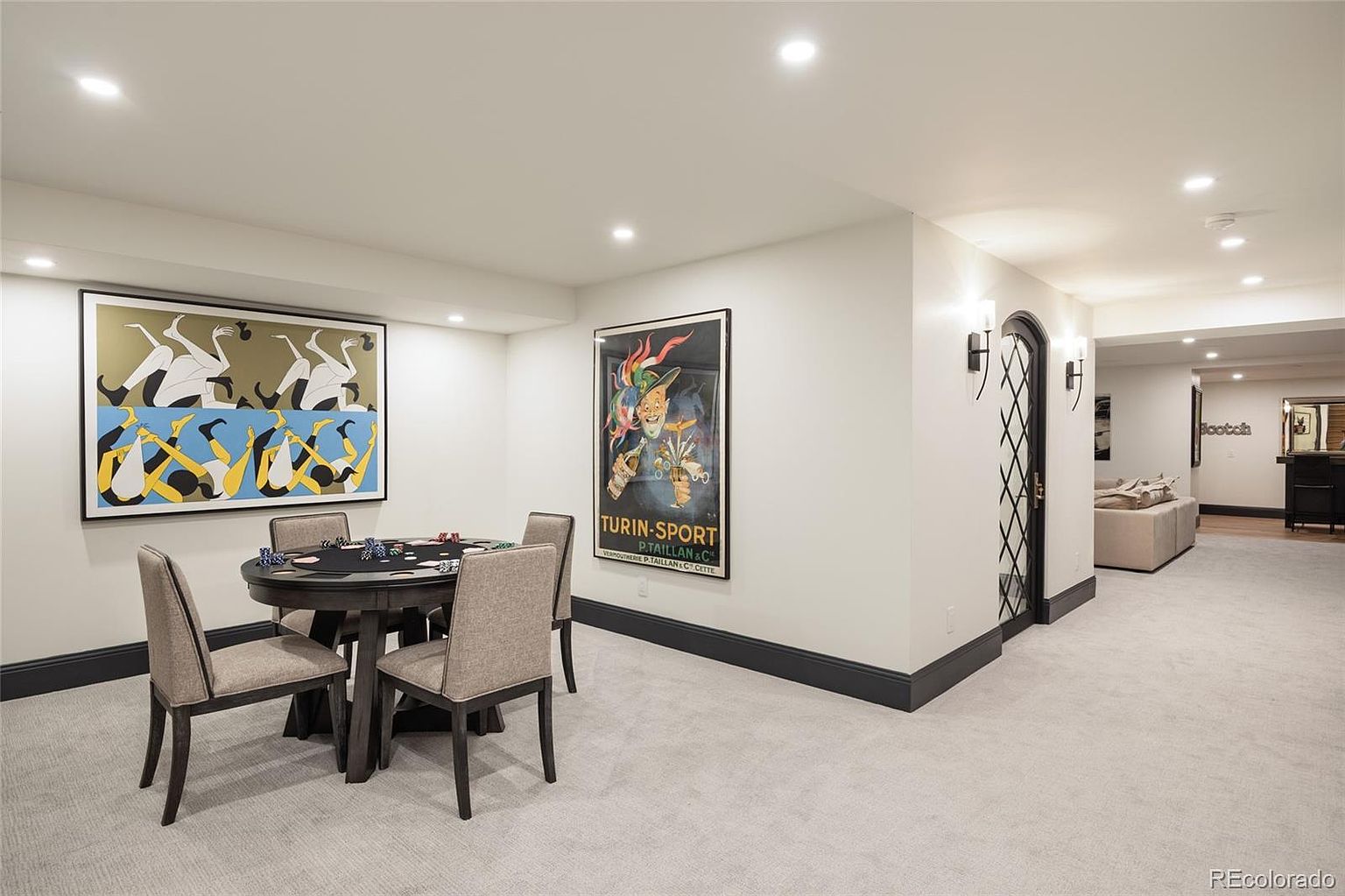 This is an interior shot of a finished basement area, featuring a poker table with chairs, artwork on the walls, and recessed lighting. The space is carpeted and has dark trim along the baseboards, creating a cozy and inviting atmosphere. An arched doorway leads to another area, suggesting a spacious and well-designed layout.