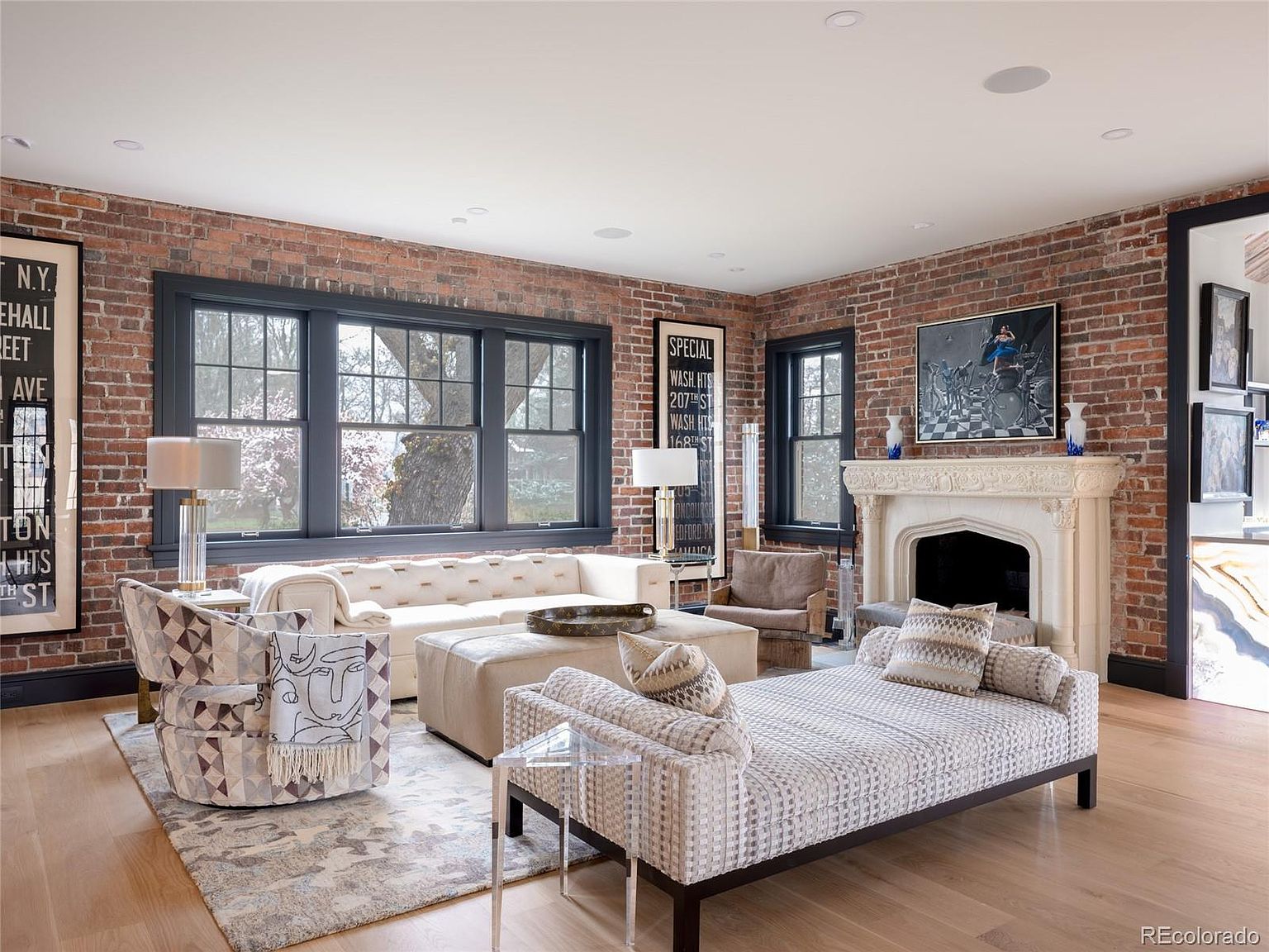 This is an interior shot of a living room featuring exposed brick walls, large windows, and a fireplace. The room is furnished with a light-colored sofa, a chaise lounge, and patterned armchairs, creating a comfortable and stylish space. The hardwood floors and neutral color palette contribute to a warm and inviting atmosphere.