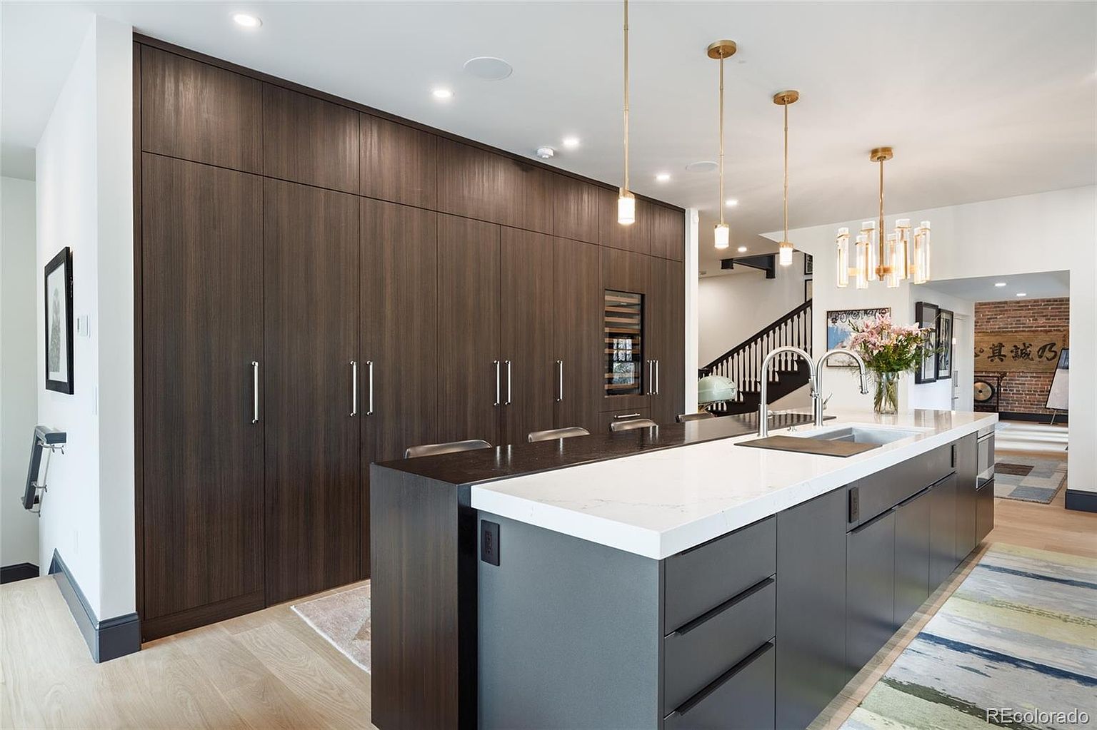 This is a modern kitchen featuring dark wood cabinetry and a large island with a white countertop. The island has gray cabinets and drawers, and there are pendant lights hanging above it. The kitchen also has a wine fridge and stainless steel appliances, with a view into the next room with stairs.