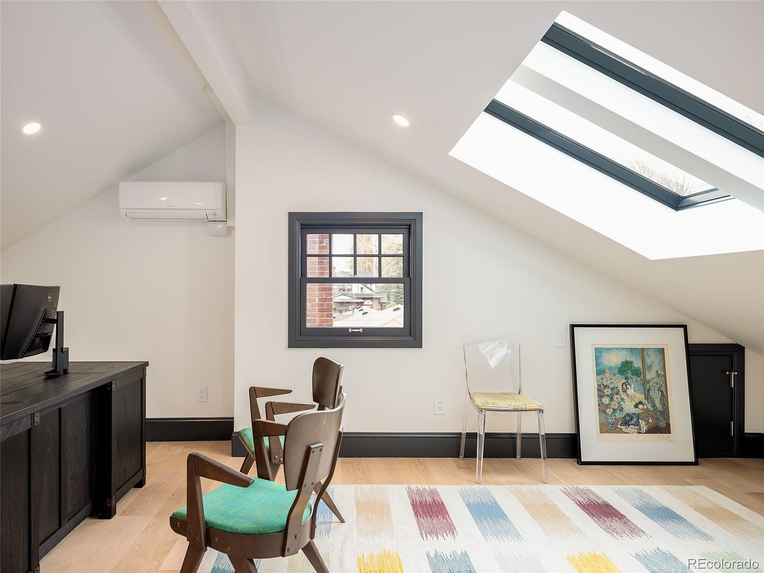 This is an interior shot of a modern office or study space, featuring a desk with a computer, two chairs, and a colorful striped rug. The room has a window and a skylight, providing ample natural light, and the walls are painted in a neutral tone, creating a bright and airy atmosphere.