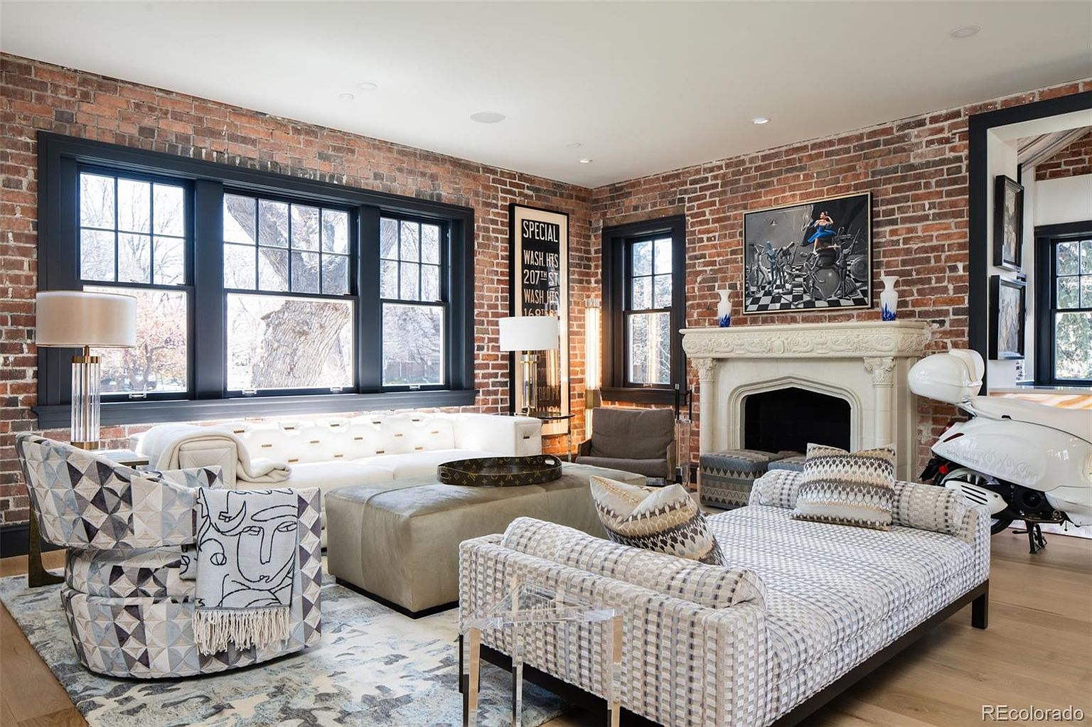 This is an interior shot of a living room featuring exposed brick walls and dark-framed windows. The room is furnished with a white sectional sofa, patterned armchairs, and a large ottoman. A fireplace with a decorative mantel adds a focal point, and a white scooter is parked near the fireplace, adding a unique touch.