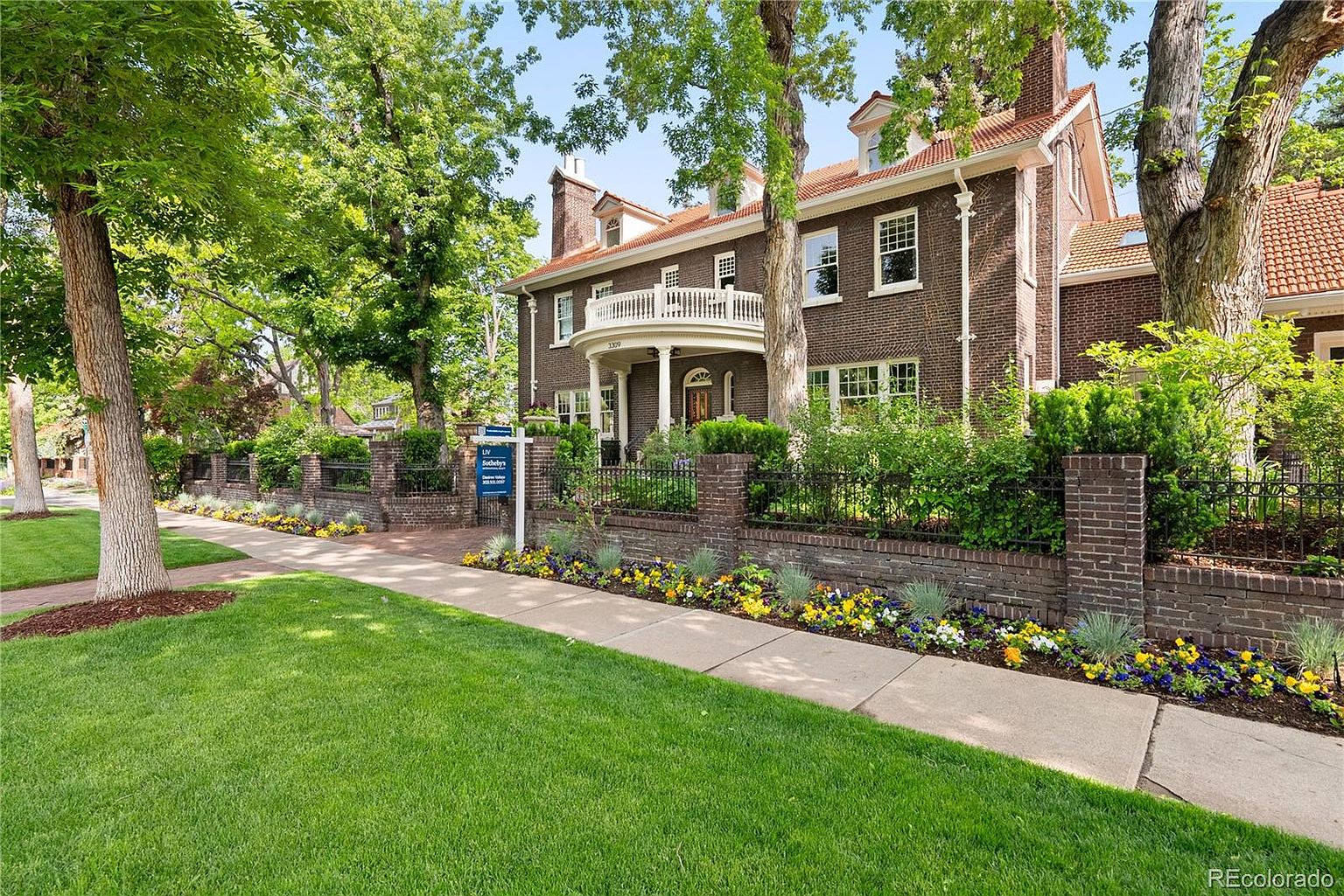 This is a front exterior view of a stately brick home with a red tile roof and a white balcony. The property features a well-manicured lawn, mature trees, and a brick fence with wrought iron accents, enhancing its curb appeal. A 'For Sale' sign is visible, indicating the property is on the market.