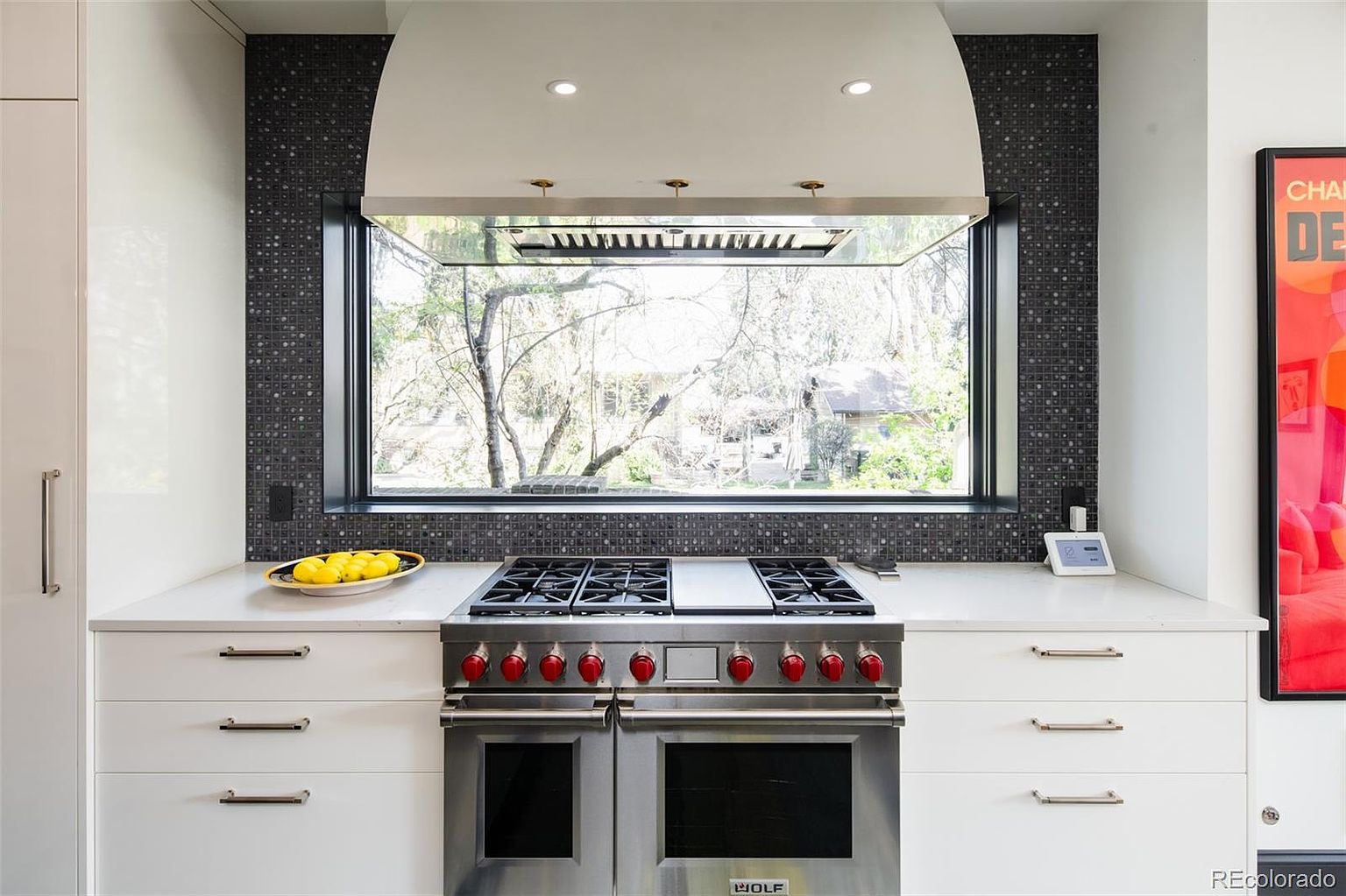 This is a well-lit kitchen featuring a stainless steel Wolf range with red knobs, set against a backdrop of dark mosaic tile and a large window offering a view of the outdoors. The white cabinetry with sleek hardware complements the modern design, while a bowl of lemons adds a touch of color to the countertop. The overall impression is clean, contemporary, and functional.