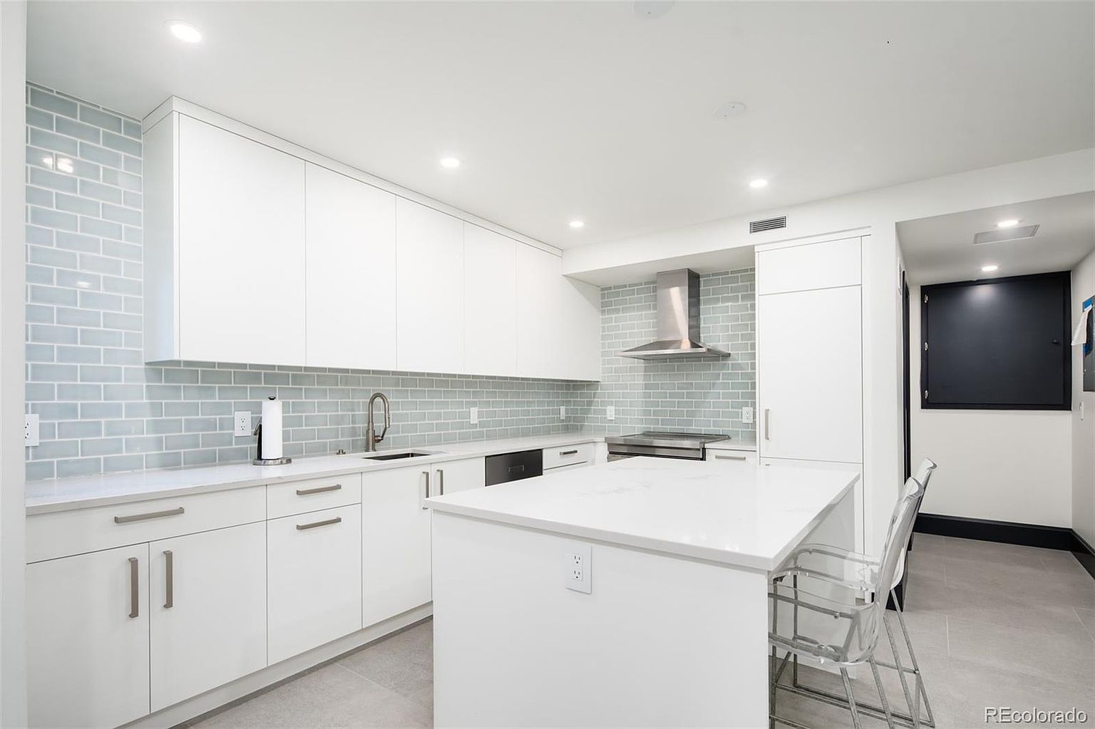 This is a bright, modern kitchen featuring sleek white cabinetry, light blue subway tile backsplash, and a large white island with seating. Stainless steel appliances and minimalist hardware contribute to the clean, contemporary aesthetic. The kitchen is well-lit with recessed lighting, creating an inviting and functional space.