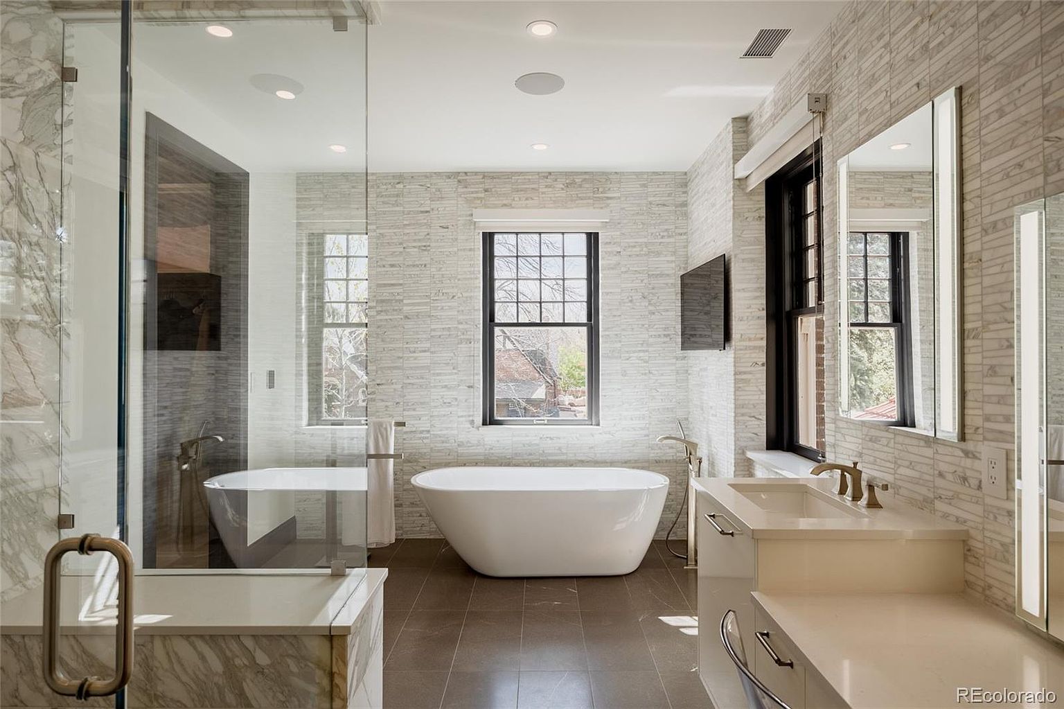 This is a luxurious primary bathroom featuring a freestanding soaking tub positioned beneath a large window, providing natural light. The space includes a glass-enclosed shower, a double vanity with modern fixtures, and elegant tile work throughout. The overall design is sophisticated and spa-like, creating a relaxing atmosphere.