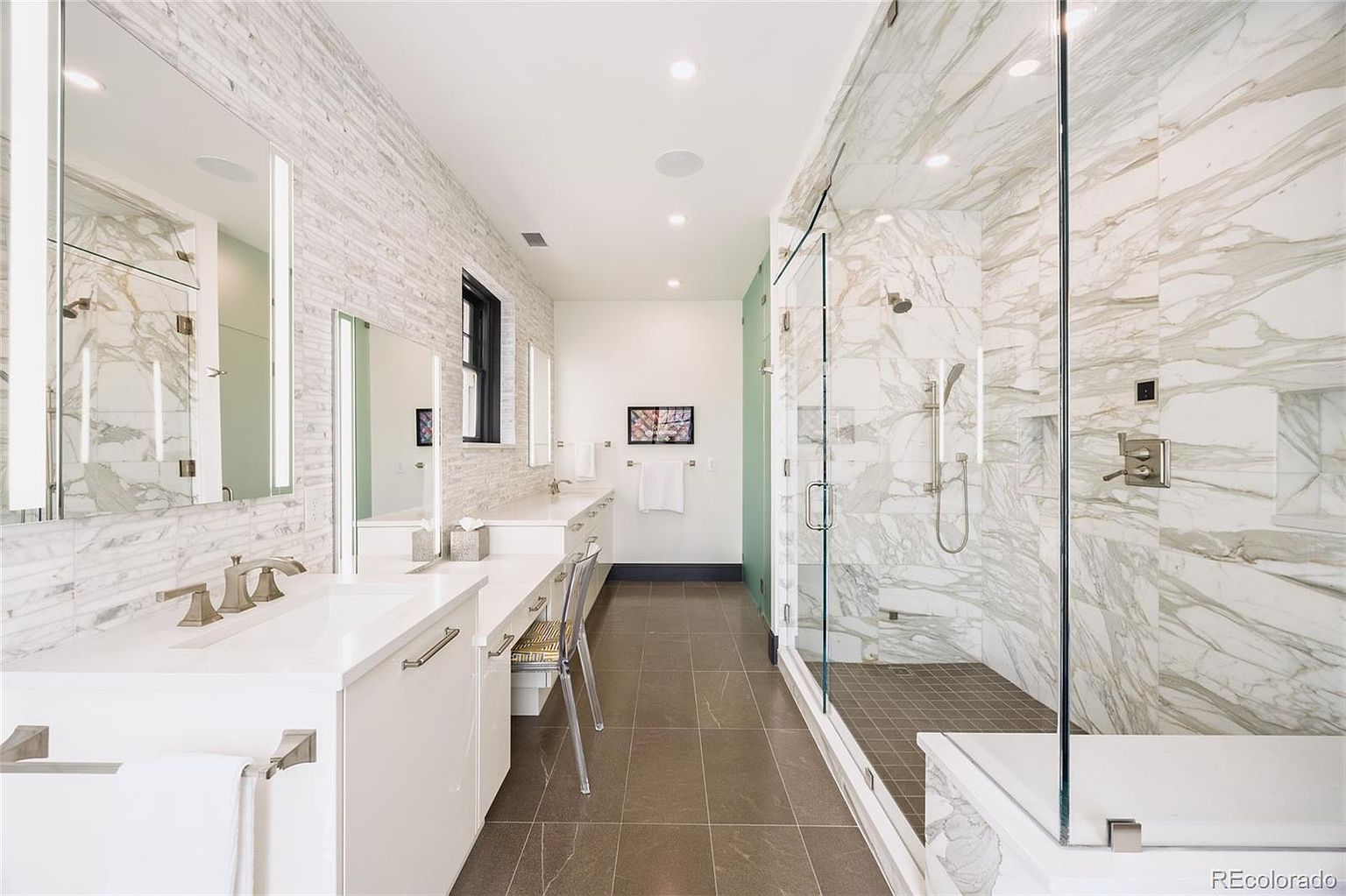 This is a luxurious primary bathroom featuring a long double vanity with white countertops and modern fixtures. A spacious walk-in shower with marble tiling and glass enclosure is visible on the right. The flooring is a neutral tile, and the overall design is sleek and contemporary, creating a spa-like atmosphere.