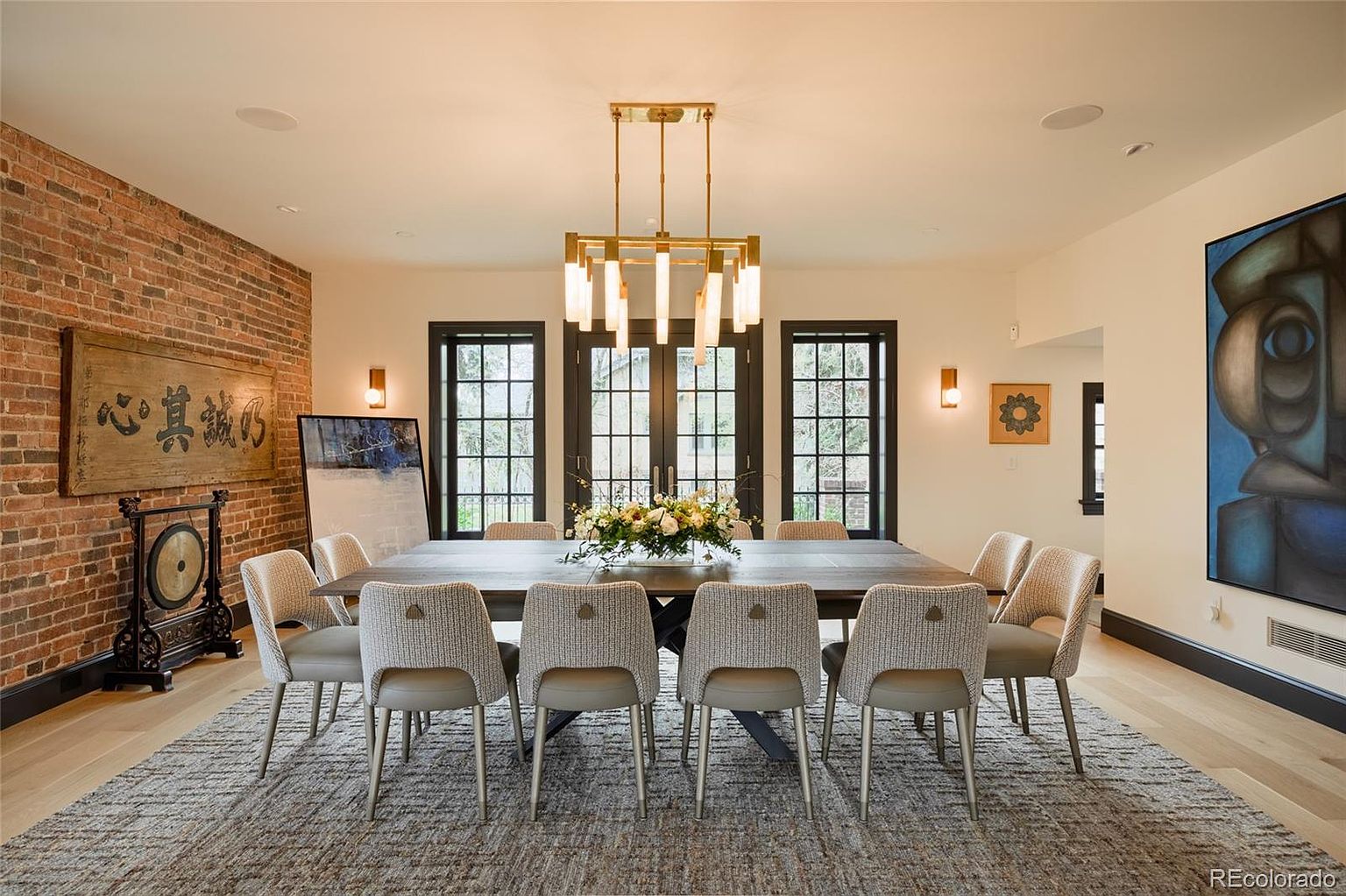 This is an interior shot of a dining room featuring a large wooden table surrounded by upholstered chairs. A modern chandelier hangs above the table, and the room is decorated with artwork and a brick accent wall. The space is well-lit and appears elegant and inviting.