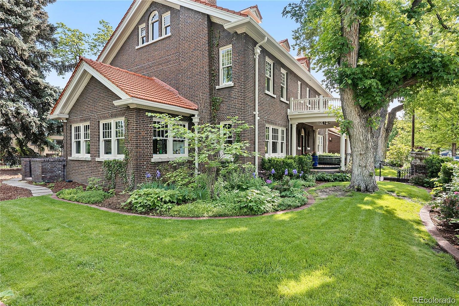 This image showcases the front exterior of a stately brick home with a well-manicured lawn and lush landscaping. The house features a red tile roof, white-framed windows, and a charming balcony. The overall impression is one of classic elegance and curb appeal, perfect for attracting potential buyers.