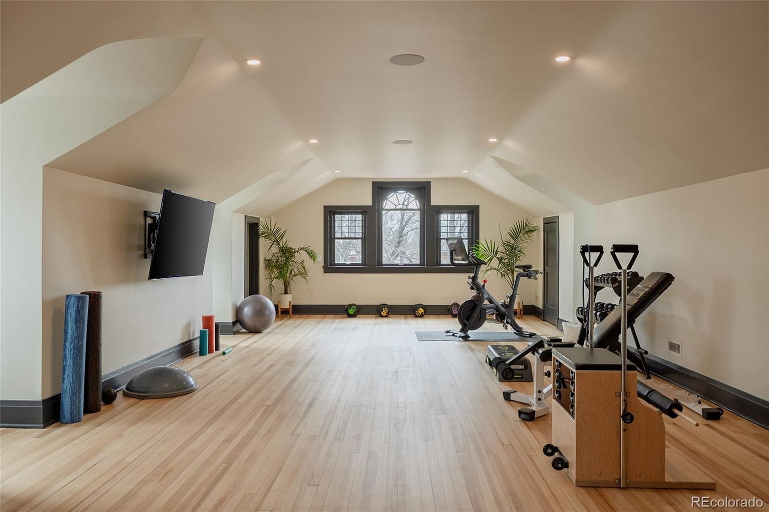 This is a spacious home gym featuring hardwood floors, neutral-colored walls, and dark trim. The room is well-lit with recessed lighting and natural light from the windows. Exercise equipment includes a stationary bike, weight bench, and various free weights, creating a dedicated space for fitness.