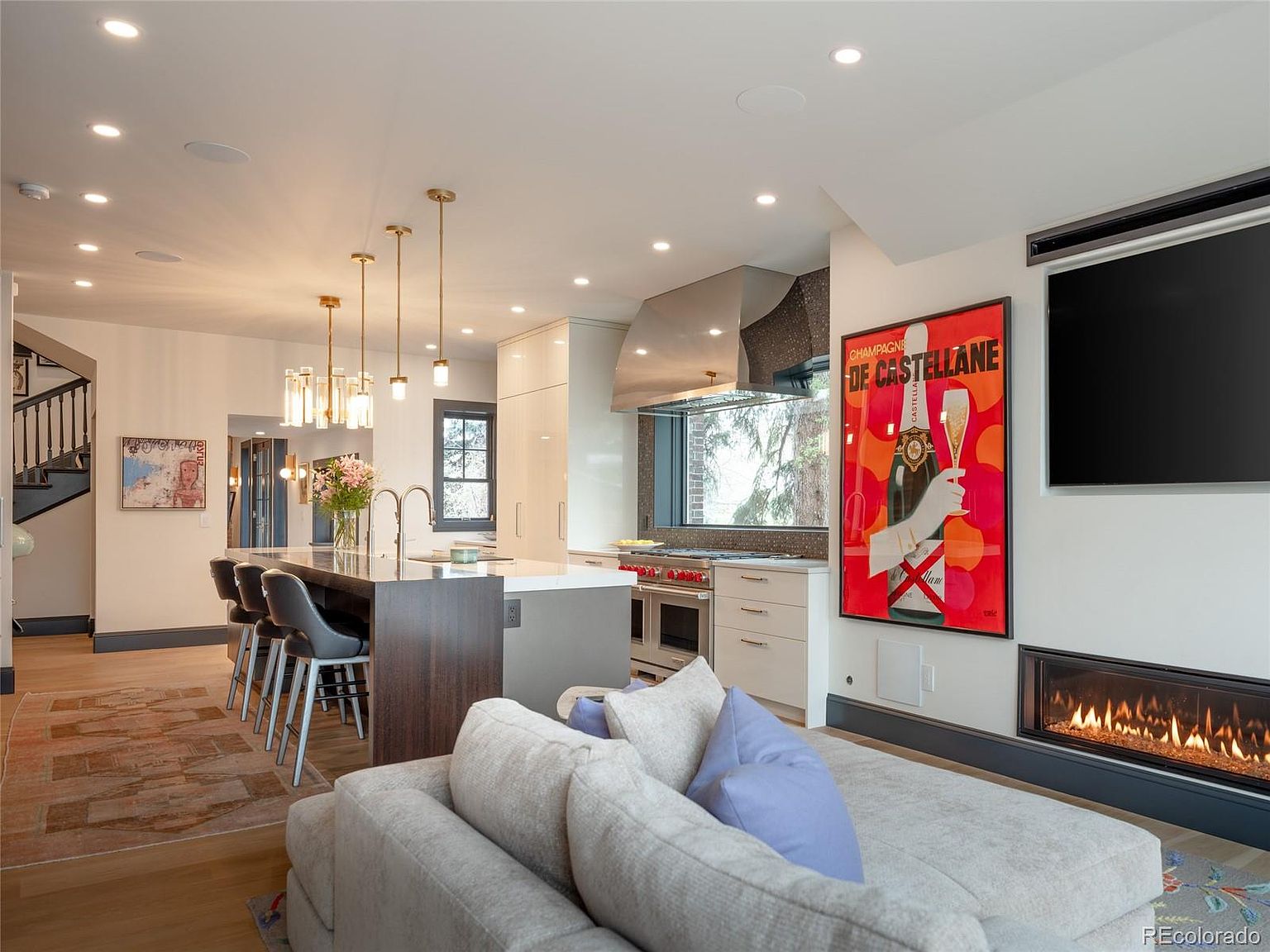 This interior shot showcases a modern kitchen and living area. The kitchen features a large island with bar seating, stainless steel appliances, and custom cabinetry, while the adjacent living area includes a fireplace and a large television. The open-concept design creates a seamless flow between the spaces, enhanced by recessed lighting and stylish decor.