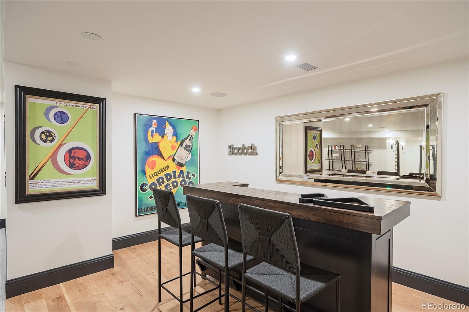 This interior shot showcases a stylish basement bar area, complete with a dark wood bar, three woven bar stools, and a mirrored wall that enhances the sense of space. The walls are adorned with vintage-style posters, adding character to the room, while the light wood flooring provides a warm contrast to the darker elements.