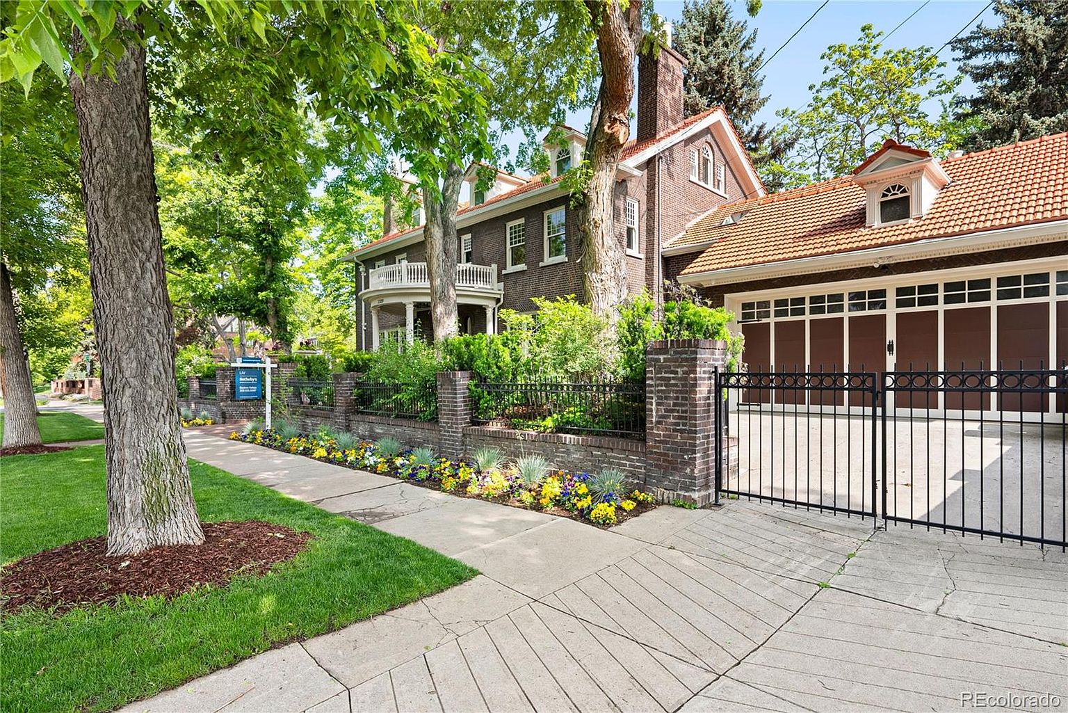 This is a front exterior view of a stately brick home with a well-manicured lawn and garden. The property features a brick fence with wrought iron accents, mature trees providing shade, and a multi-car garage with a tiled roof. The overall impression is one of classic elegance and curb appeal.