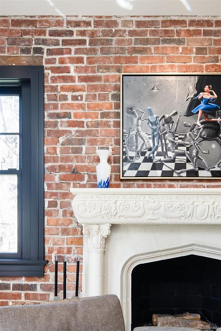 This interior shot showcases a living room with a prominent brick wall and a dark-framed window. A white, ornate fireplace mantel displays a vase and a framed painting, adding an artistic touch. The room exudes a blend of rustic charm and sophisticated elegance.
