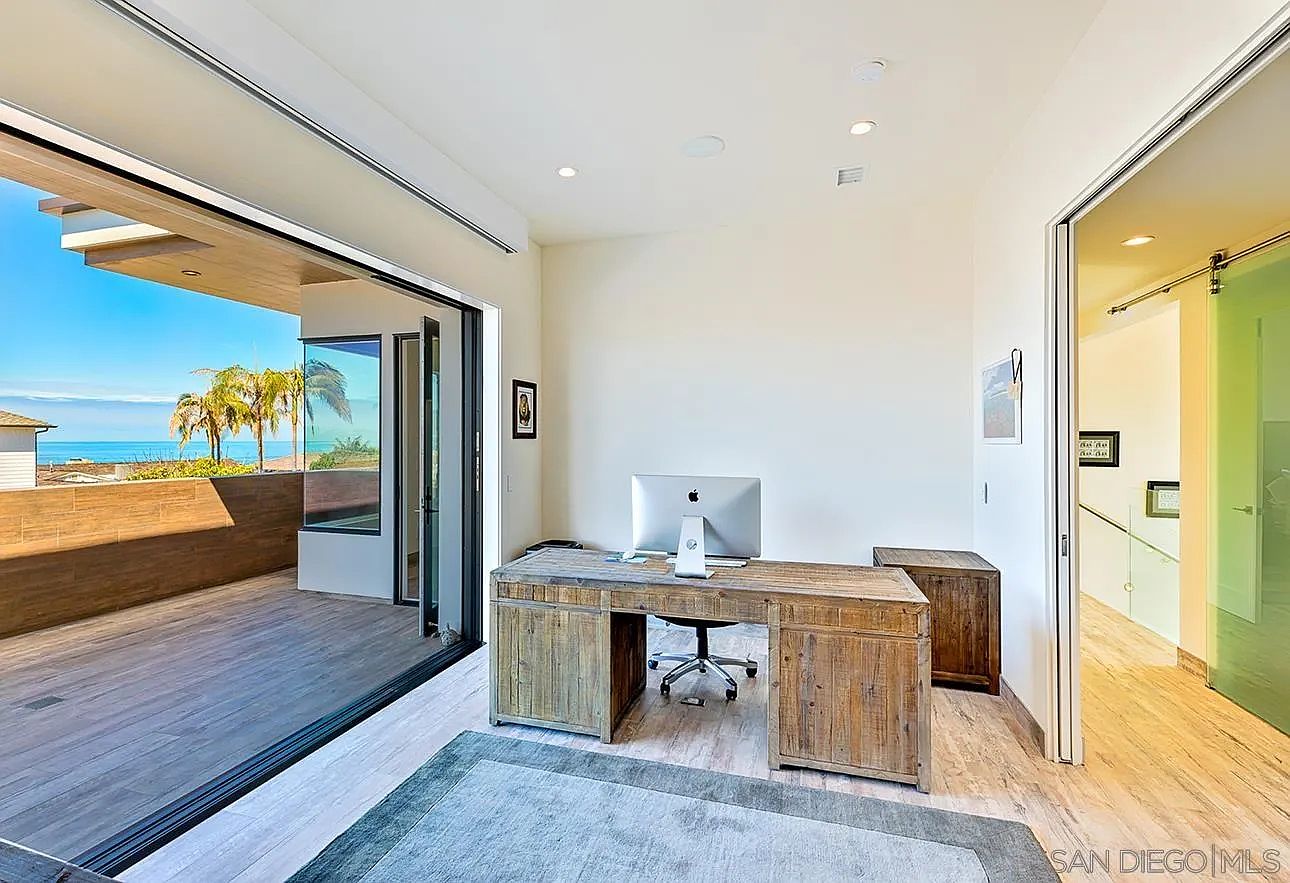 This is an interior shot of a modern office space featuring a rustic wooden desk with a desktop computer. The room has a large sliding glass door leading to a balcony with ocean views, bringing natural light into the space. The flooring is light-colored tile, and a gray rug adds texture to the room.