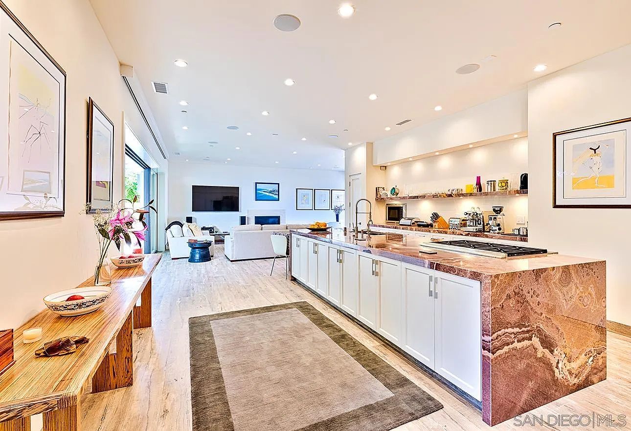 This is a bright and open kitchen and living area. The kitchen features a large island with a unique marble countertop and white cabinetry, complete with modern appliances. The living area is spacious, with a fireplace, comfortable seating, and large windows that let in plenty of natural light, creating an inviting and luxurious atmosphere.