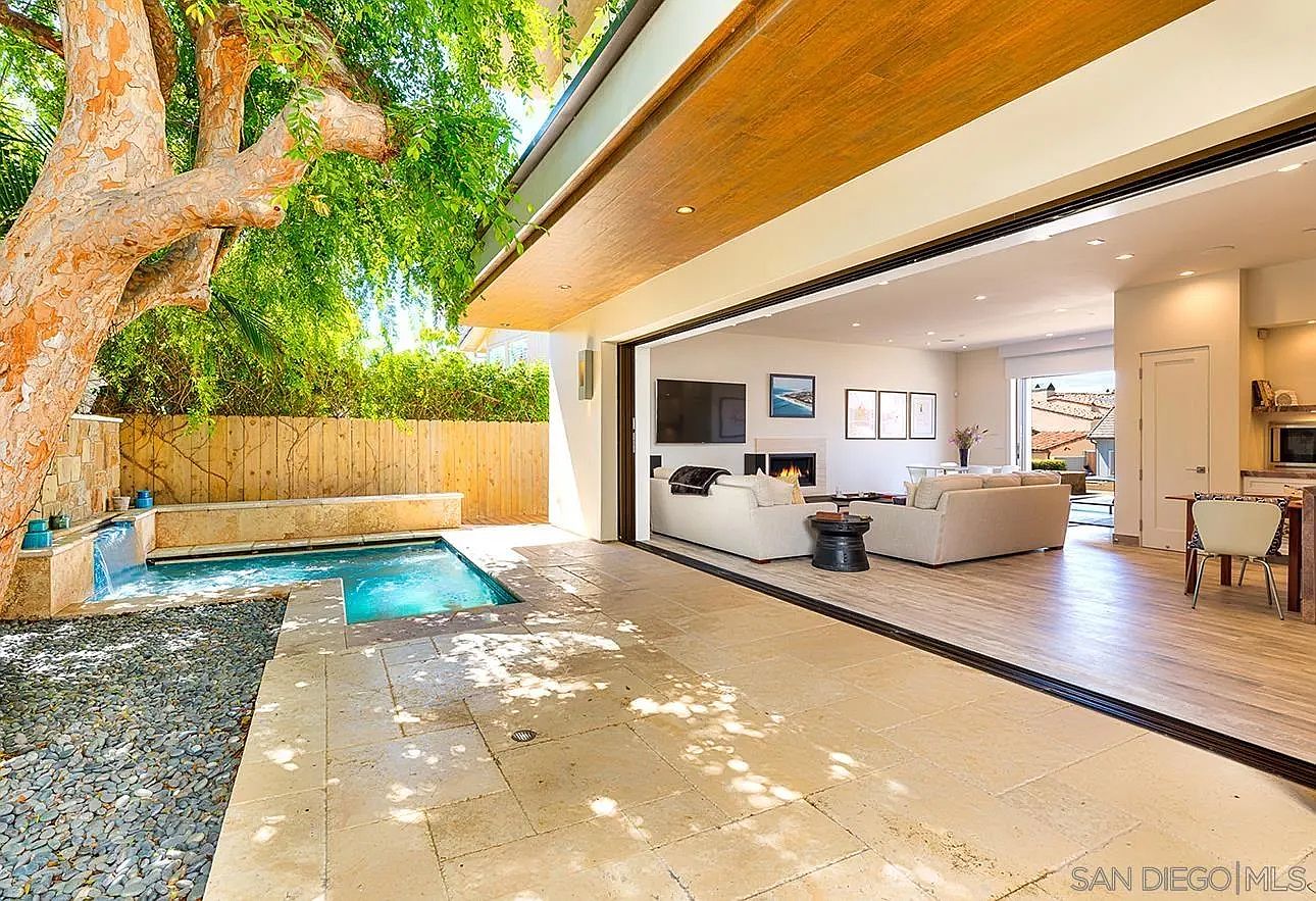 This image showcases a luxurious outdoor living space with a pool and seamless indoor-outdoor transition. The pool area features a small waterfall and is surrounded by stone pavers and lush greenery. Large sliding glass doors open to a modern living room, creating an inviting and open atmosphere perfect for entertaining or relaxation.