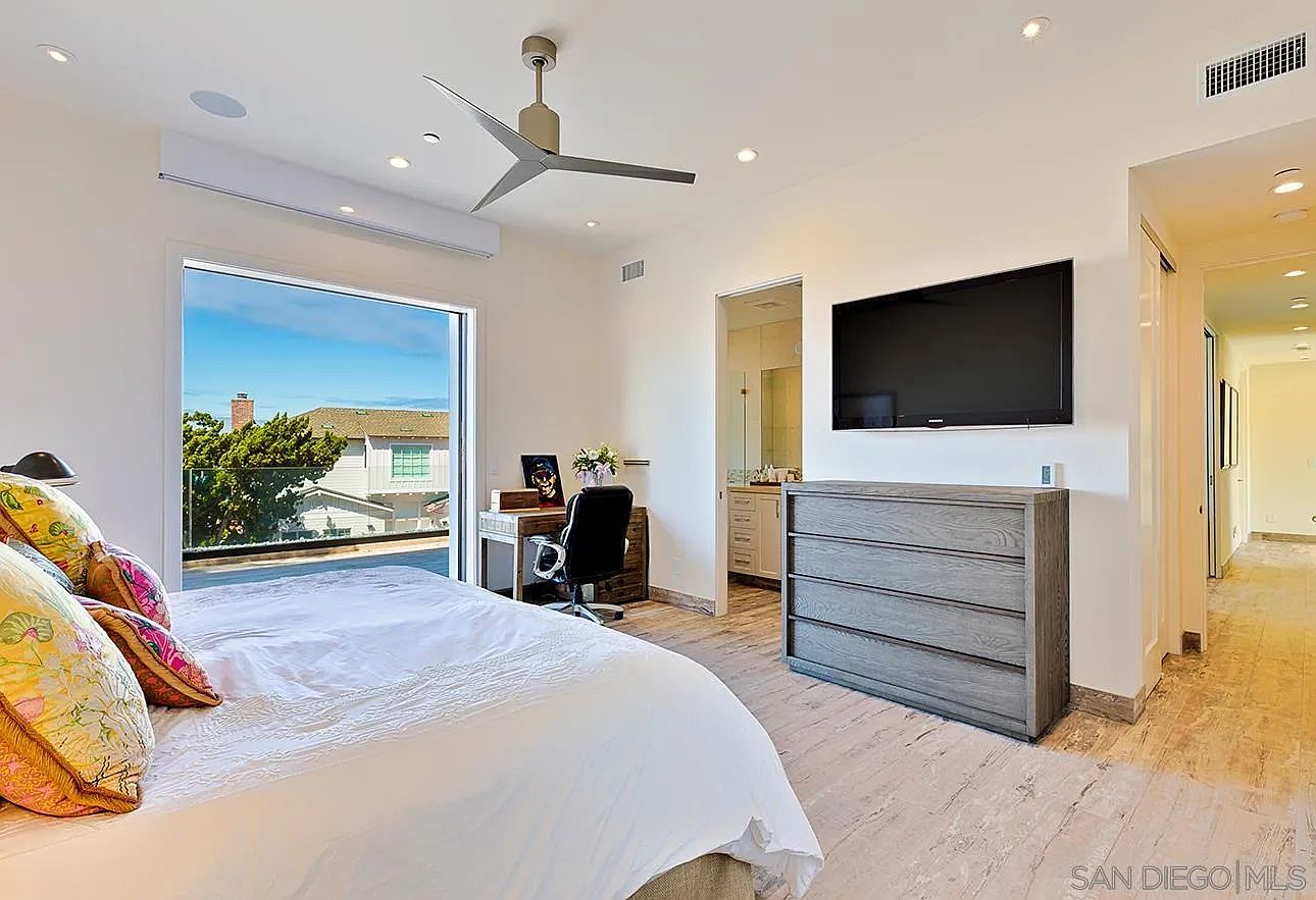 This is a bright and airy primary bedroom featuring a large window with a view, a modern ceiling fan, and a wall-mounted television above a gray dresser. A desk and chair are positioned near the window, creating a functional workspace. The room has a clean and contemporary aesthetic with light-colored flooring and walls.