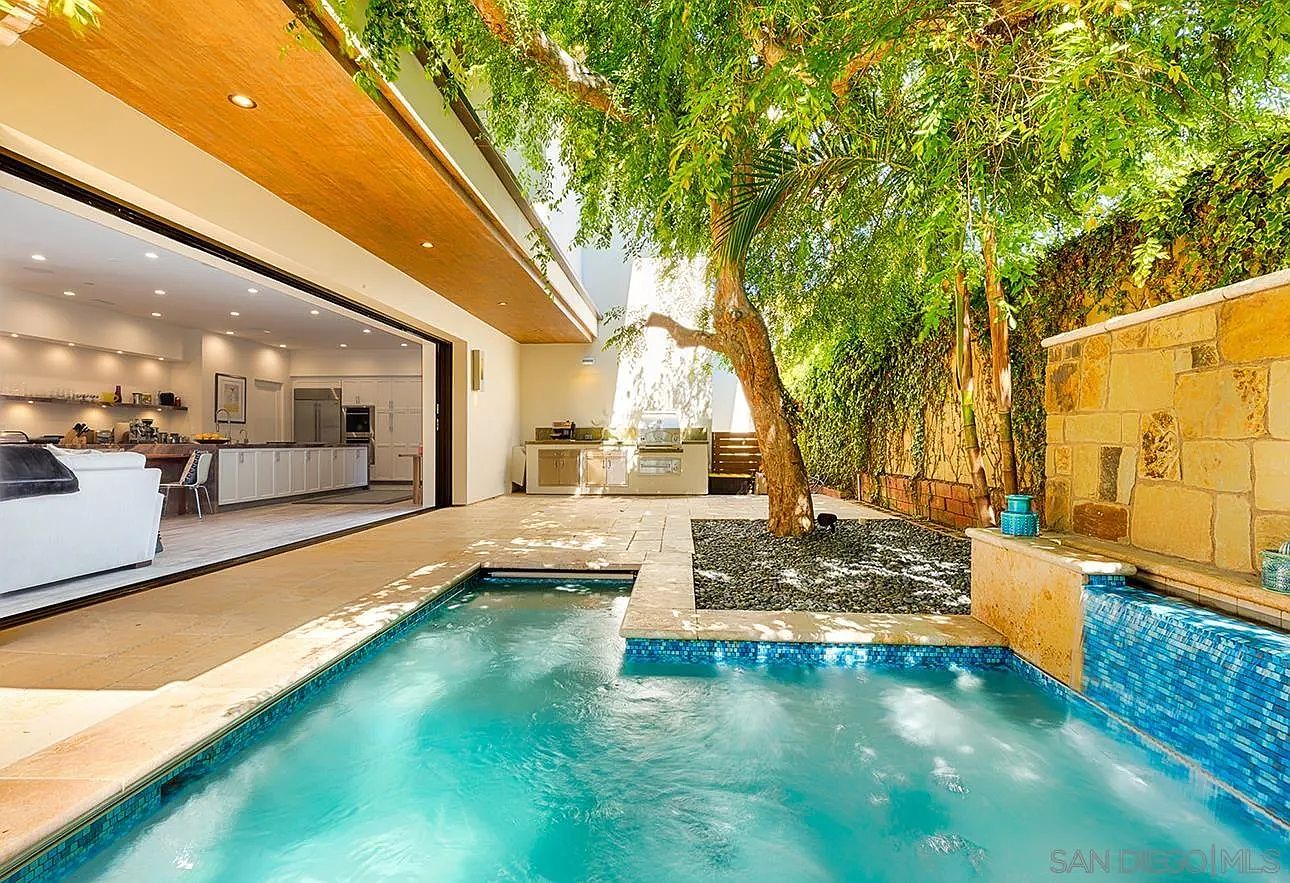 This image showcases a luxurious outdoor pool and patio area, seamlessly blending indoor and outdoor living. The pool features a modern design with blue mosaic tiling and a small waterfall feature. Adjacent to the pool is a well-manicured garden area with a mature tree, providing shade and privacy, while the open doorway leads into a bright, modern kitchen, creating an inviting and cohesive living space.