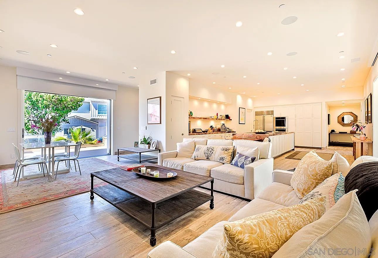 This is an interior shot of a spacious living room that seamlessly transitions into a dining area and kitchen. The living room features comfortable sofas adorned with decorative pillows, a rustic coffee table, and large windows that provide ample natural light and views of the outdoors. The open floor plan creates an inviting and airy atmosphere, perfect for modern living.