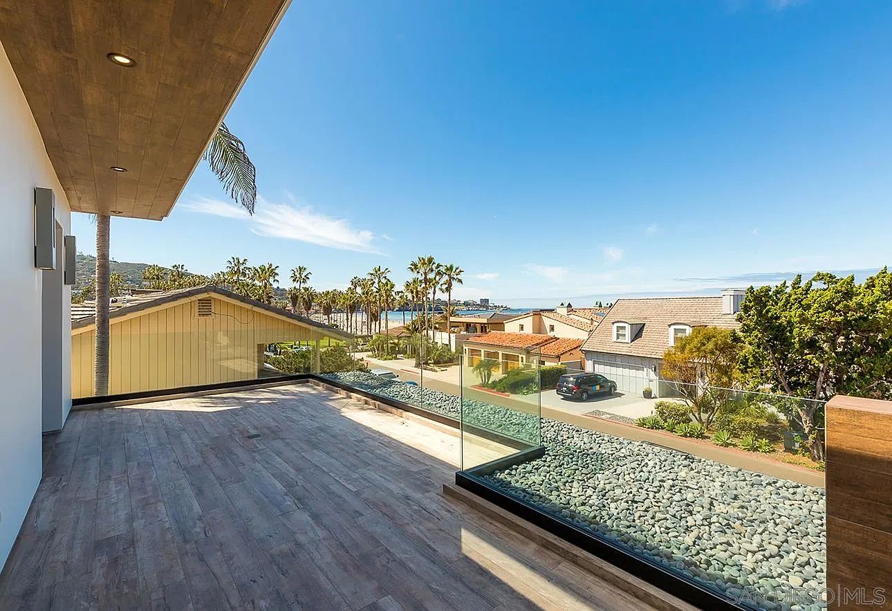 This image showcases a spacious balcony with a wooden deck and a modern glass railing filled with decorative stones. The view extends over residential houses towards the ocean, framed by palm trees and a clear blue sky. The scene evokes a sense of luxury and relaxation, highlighting the property's desirable location and outdoor living space.