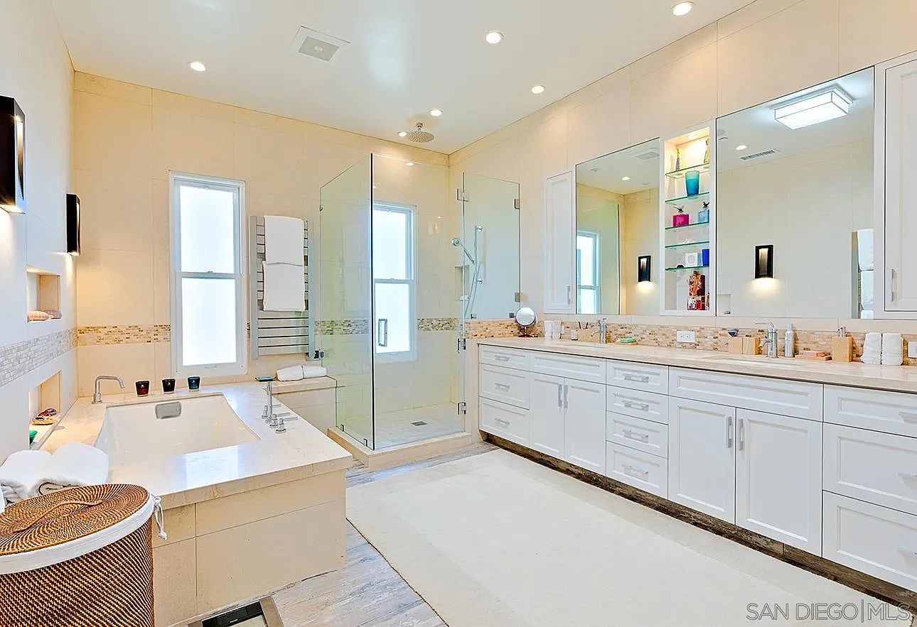 This is a spacious primary bathroom featuring a large soaking tub with a marble surround, a glass-enclosed shower, and a double vanity with white cabinetry and a light-colored countertop. The walls are tiled in a neutral tone, and recessed lighting illuminates the space. The overall impression is luxurious and spa-like.