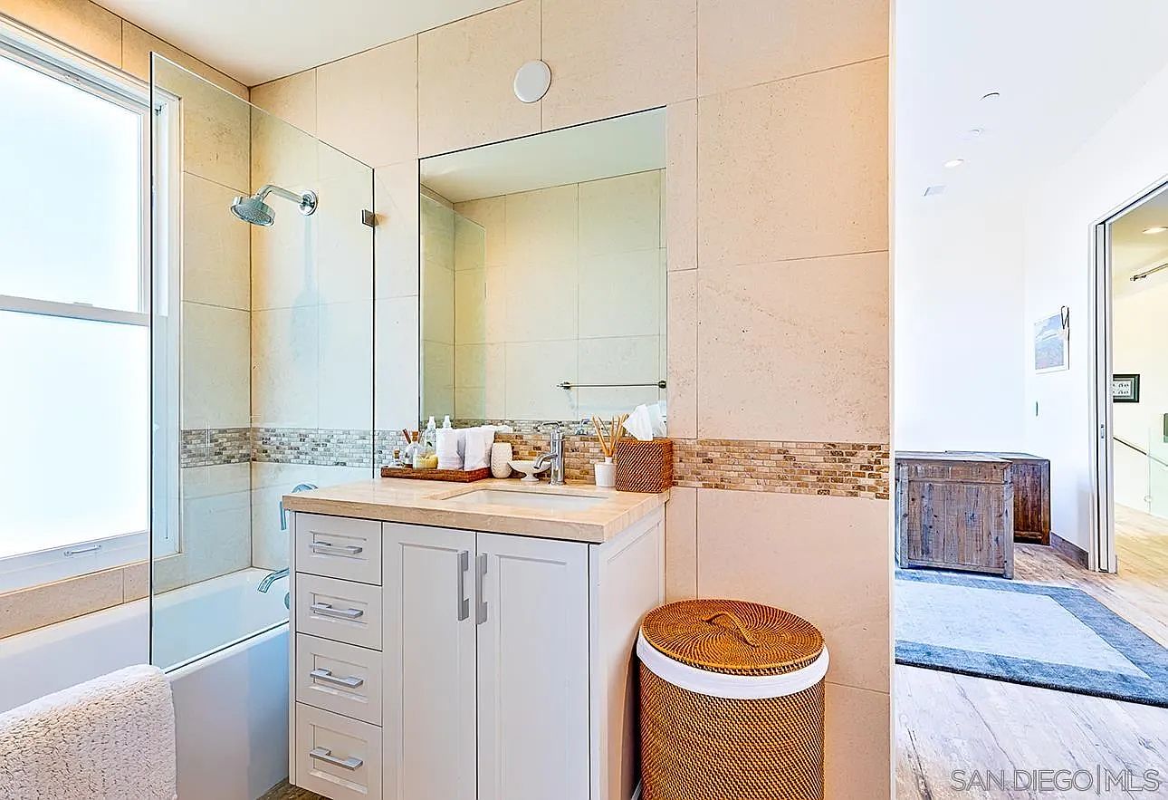 This is a well-lit bathroom featuring a vanity with a light-colored countertop and white cabinetry. A large mirror hangs above the sink, reflecting the neutral-toned tile walls. A bathtub with a glass shower screen is visible to the left, and a woven laundry basket sits to the right of the vanity, adding a touch of texture.