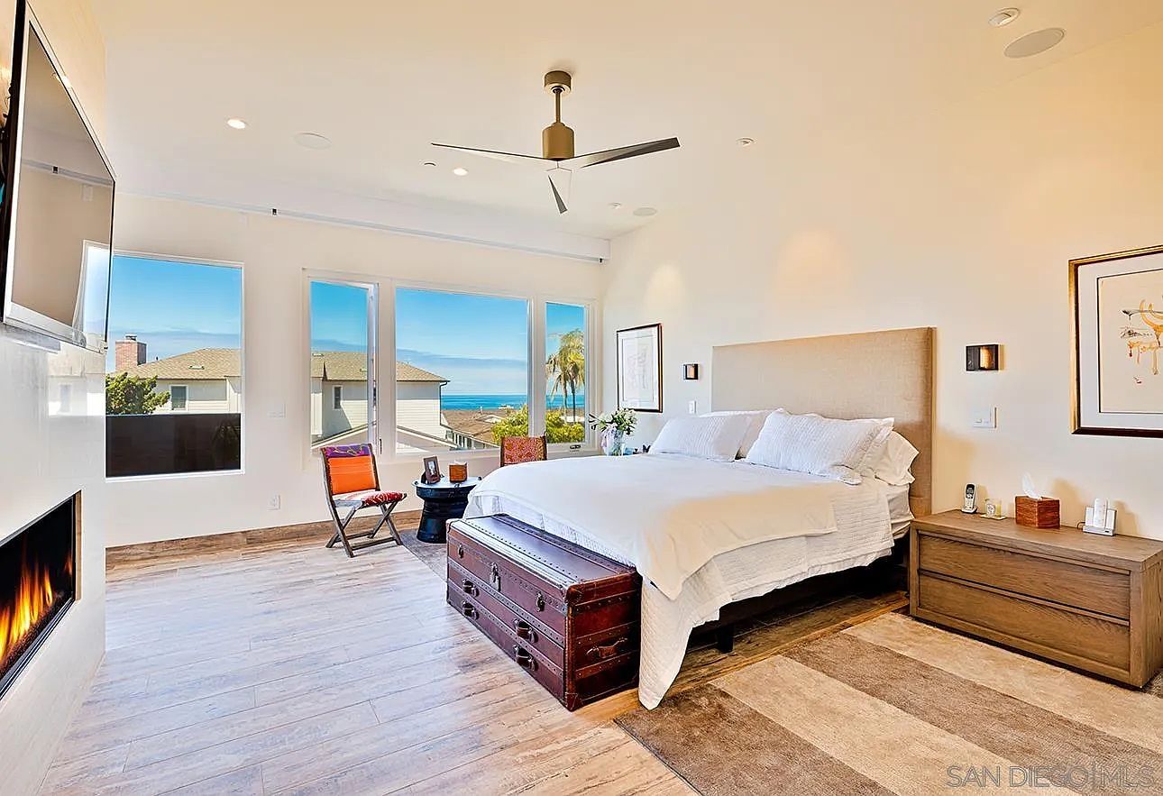 This is a bright and airy primary bedroom featuring a large bed with white linens, a wooden chest at the foot of the bed, and a matching nightstand. Large windows offer a beautiful ocean view, and a modern fireplace adds a touch of luxury. The room has a relaxed and sophisticated coastal style.